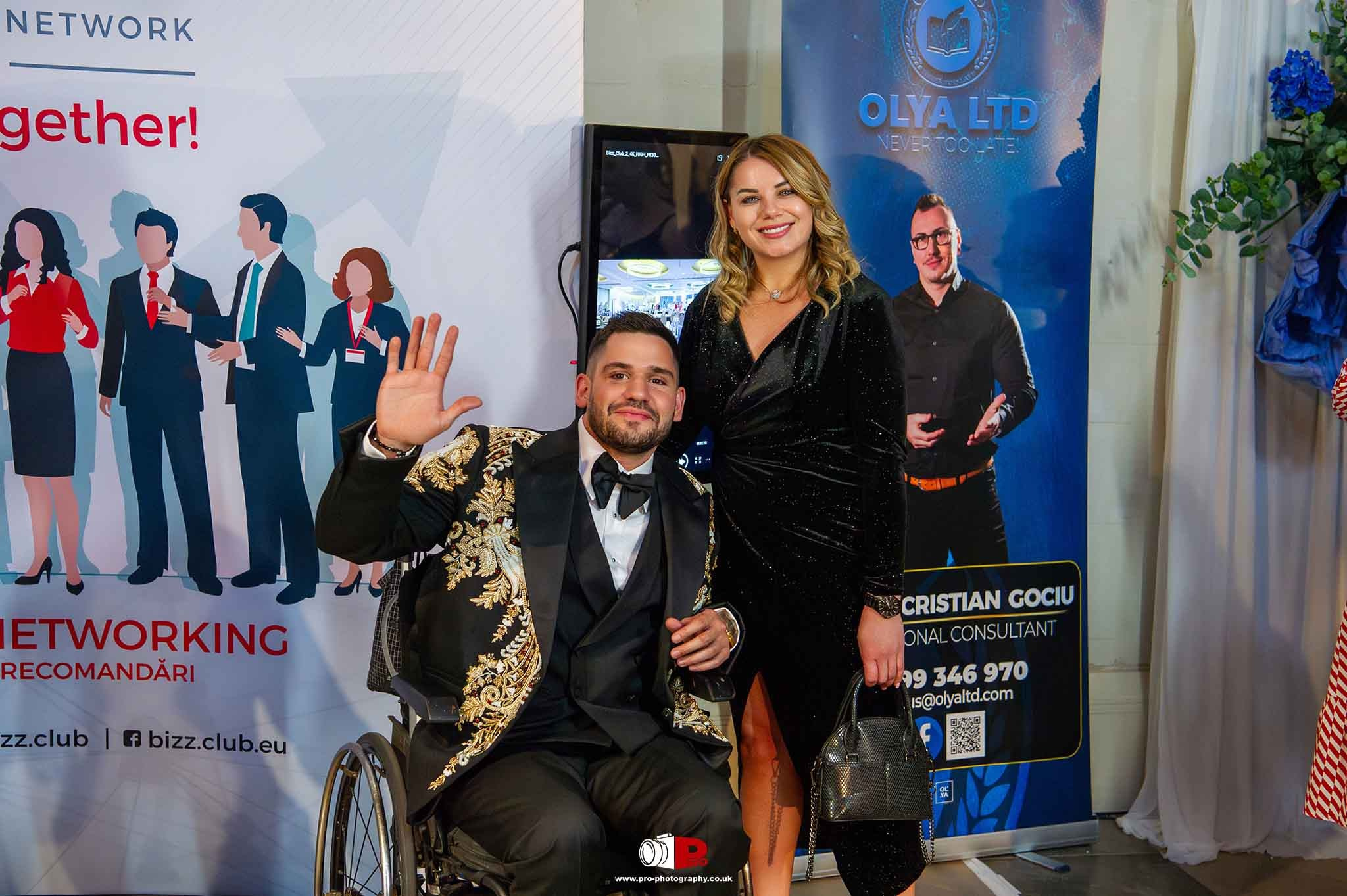 A smiling man in a wheelchair wearing a gold-embroidered black tuxedo posing next to a woman in a black evening dress at a professional networking event, standing in front of business promotional banners.