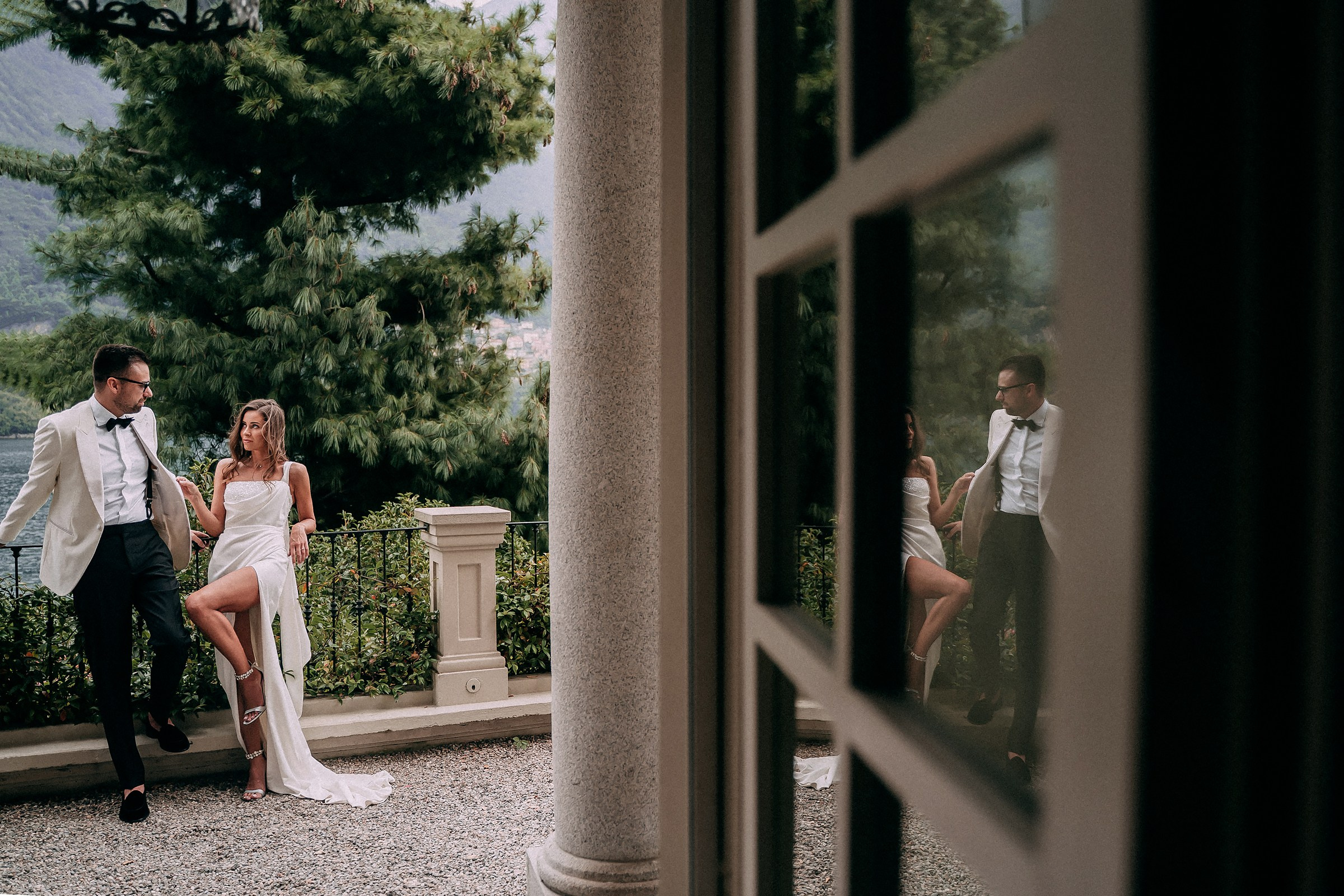 Romantic couple at scenic outdoor wedding venue. Bride in white gown with high slit stands beside groom in white jacket and black trousers near a stone column, with mountain landscape and greenery in the background. Their reflection appears in a glass window, adding depth and elegance to the moment.