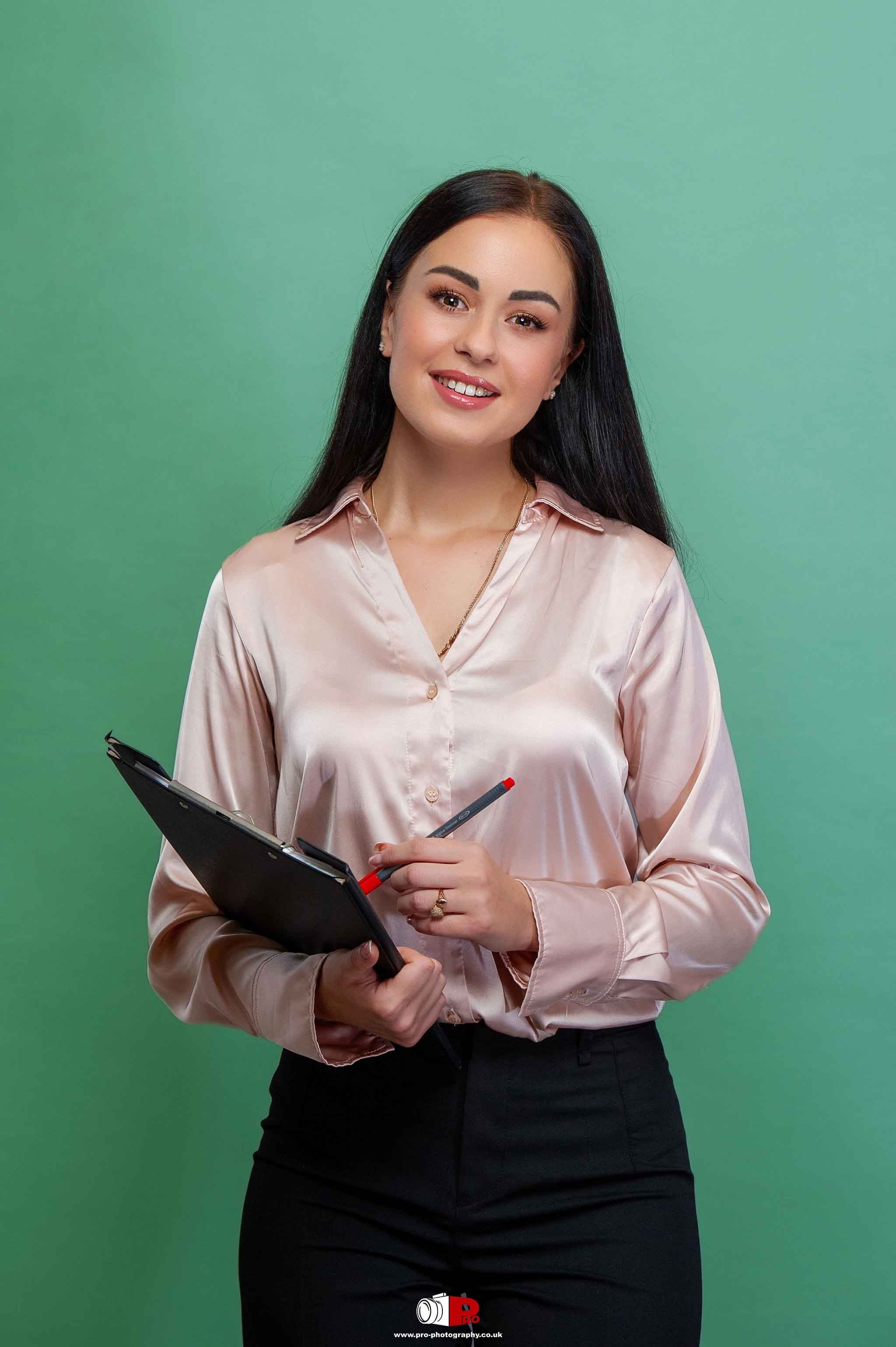 A smiling woman holding a clipboard and pen, dressed in a pink silk blouse, standing against a green background.
