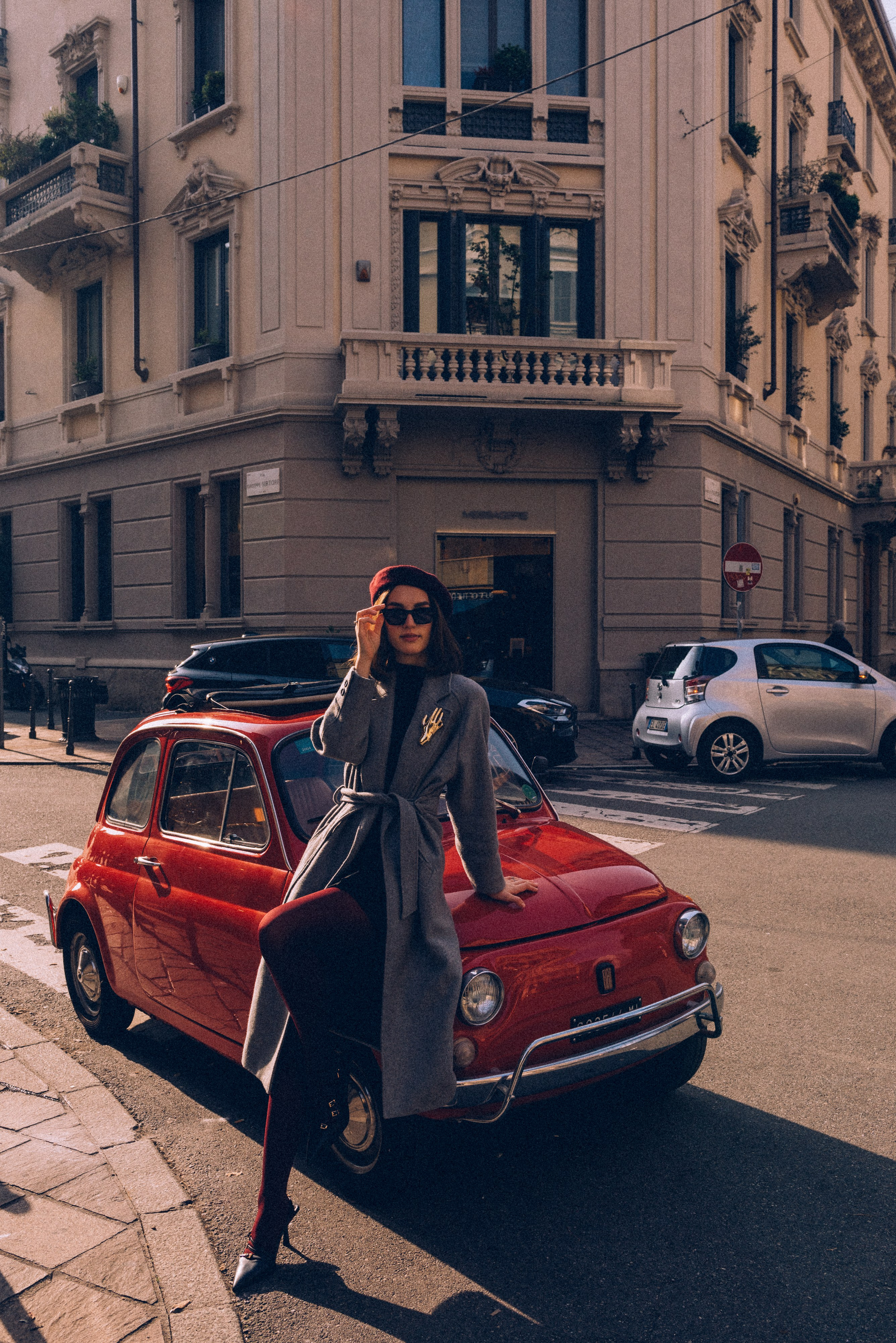 Retro charm in Milan: a woman in a chic outfit leans on a vintage red Fiat 500 in a picturesque urban setting. Milan photographer for tourists