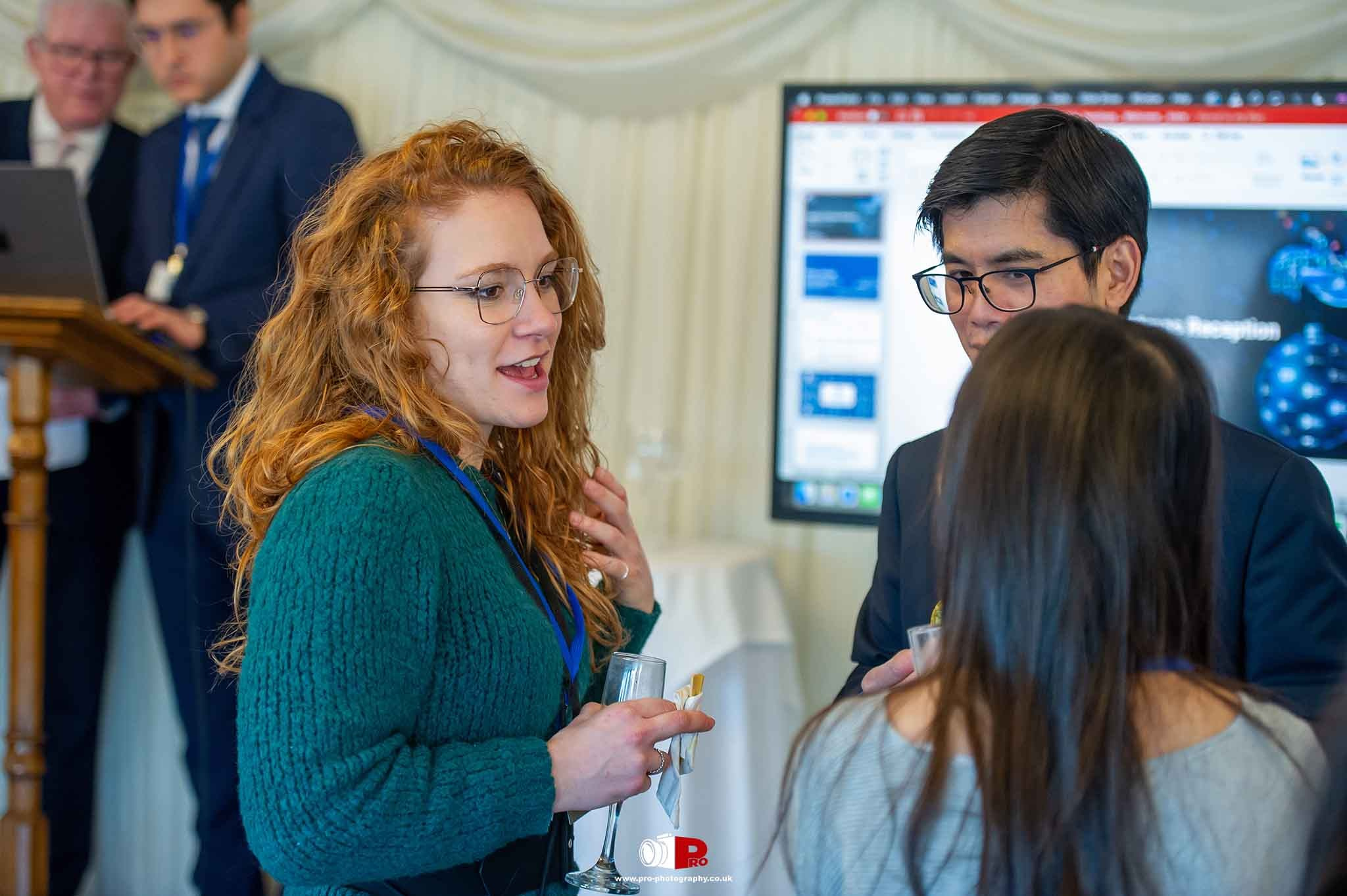 A group of professionals engaged in conversation at a networking event with presentation slides in the background.