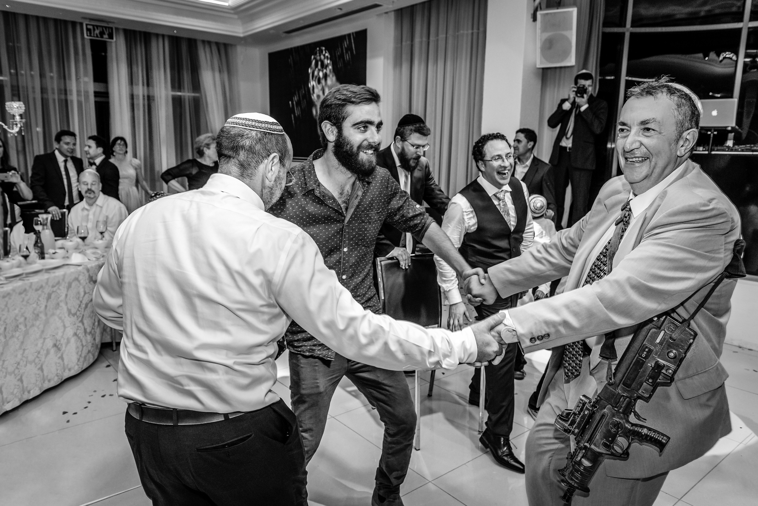 A black and white photograph captures the joyful celebration of a Jewish wedding. In the foreground, a group of men enthusiastically dance, radiating joy and energy. The men are dressed in a variety of formal attire, including suits and vests. Among them, one man stands out, dancing with a machine gun slung over his shoulder.