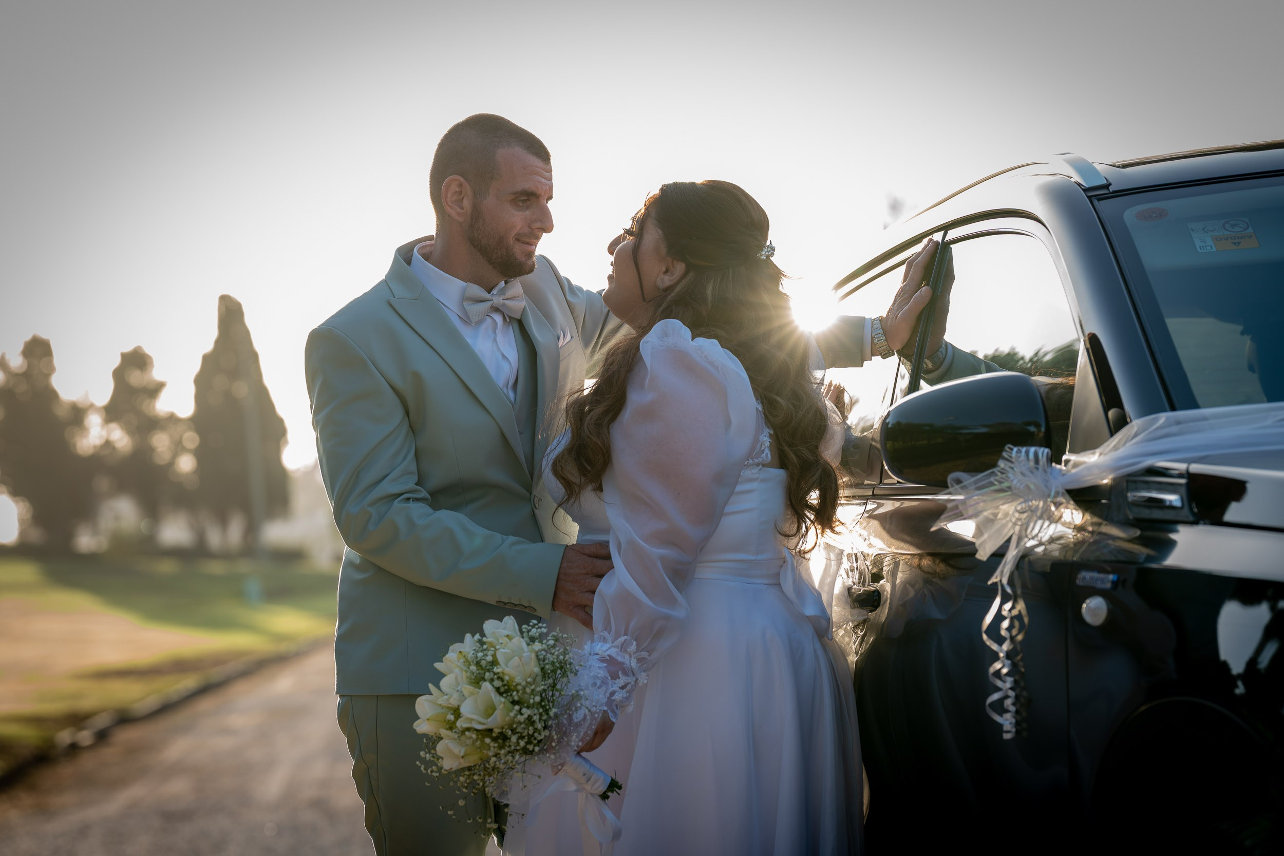 Ella and Matan share a quiet moment beside their wedding car at sunset