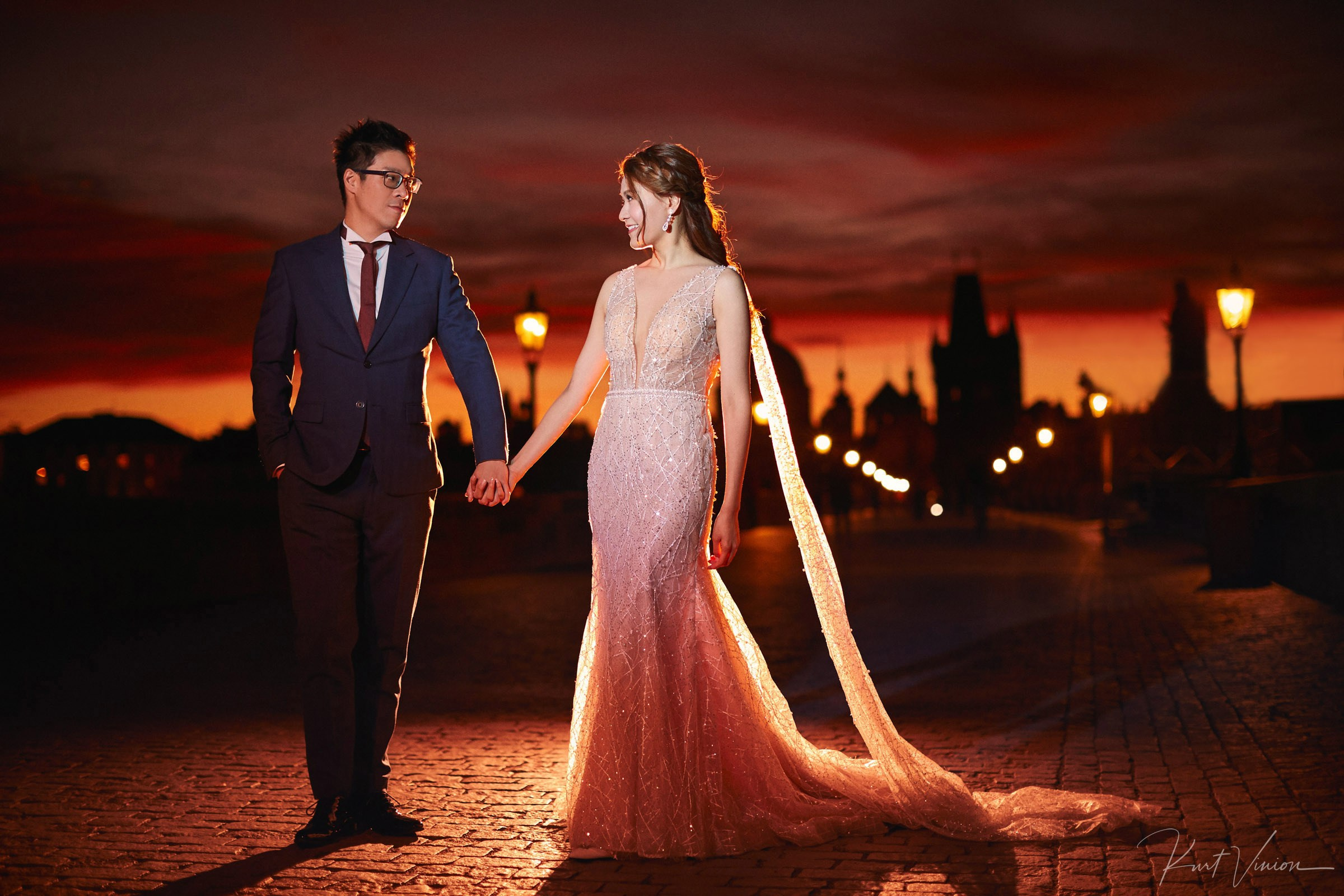 Stylish Asian couple in evening attire hold hands atop the Charles Bridge as the early morning sunlight breaks above the gothic tower behind them