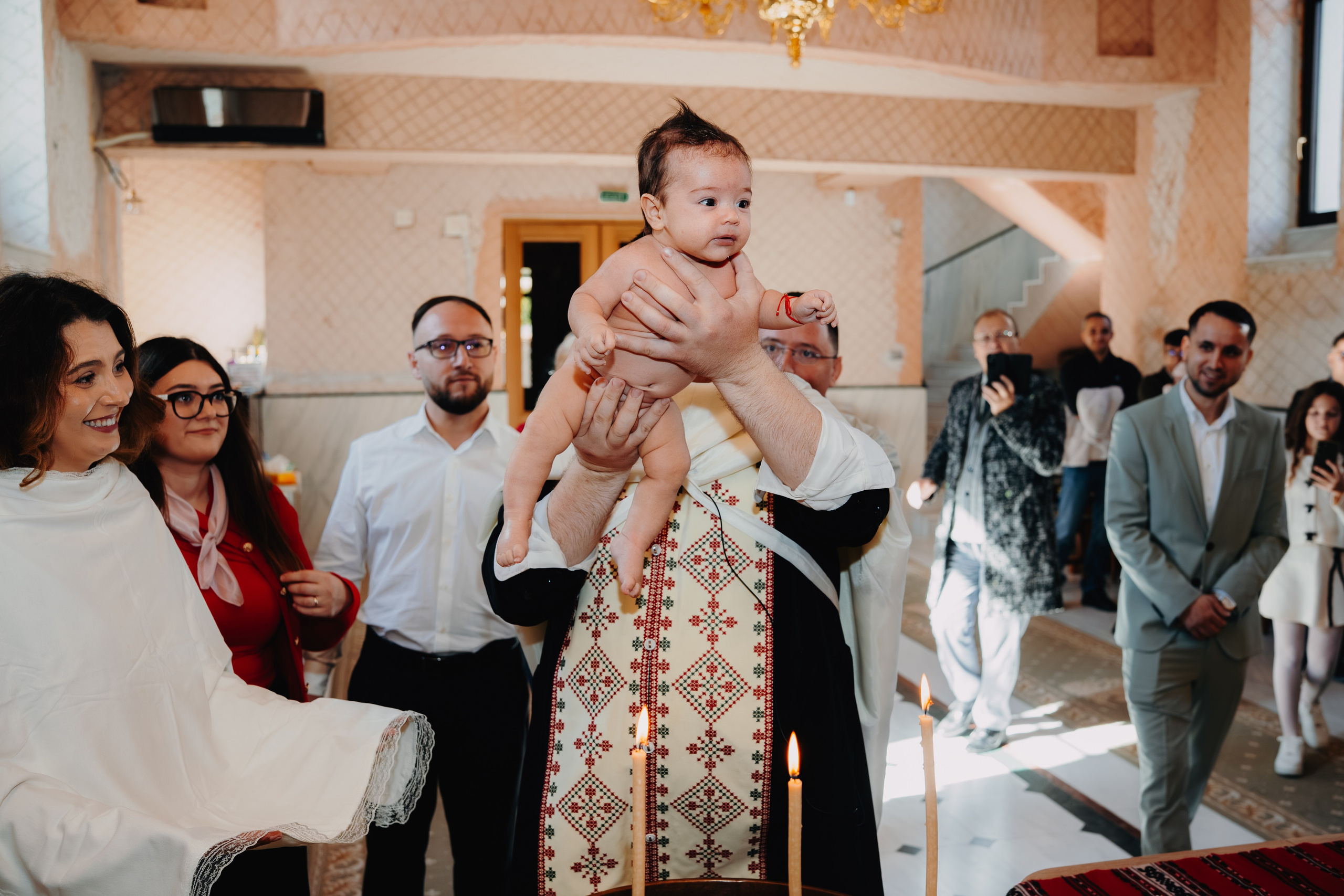 Momentul central al botezului lui Arthur Mihail în Catedrala Bârlad: preotul îl ține deasupra cristelniței, gata să-l scăldă, în timp ce familia și nașii privesc cu emoție și curiozitate solemnă din ambele părți ale altarului.