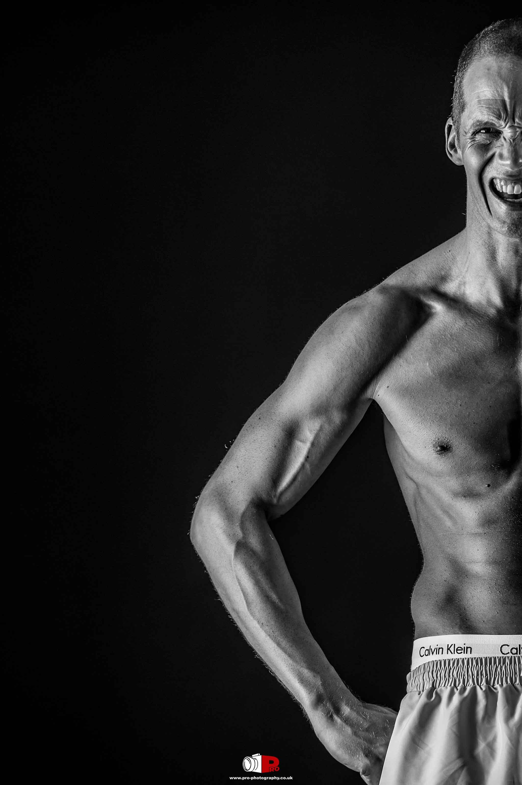 Close-up monochrome shot of a shirtless male fitness model smiling with muscular definition against a dark background.