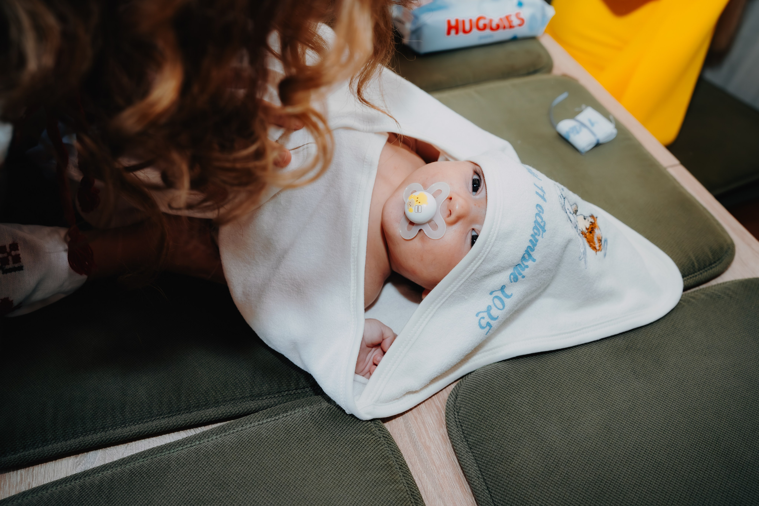 Arthur Mihail este așezat pe masa botezului, înfășat într-un prosop alb, în momentul de dinaintea scăldării sfinte. Fotografie intimă care captează inocența și vulnerabilitatea solemnă din fața ritualului.