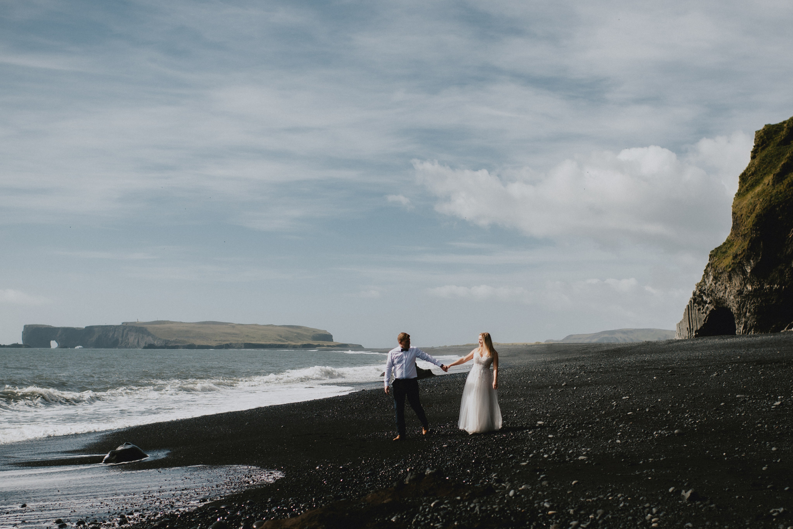 Bride and groom standing on a black sand beach in South Iceland, with dramatic waves in the background.