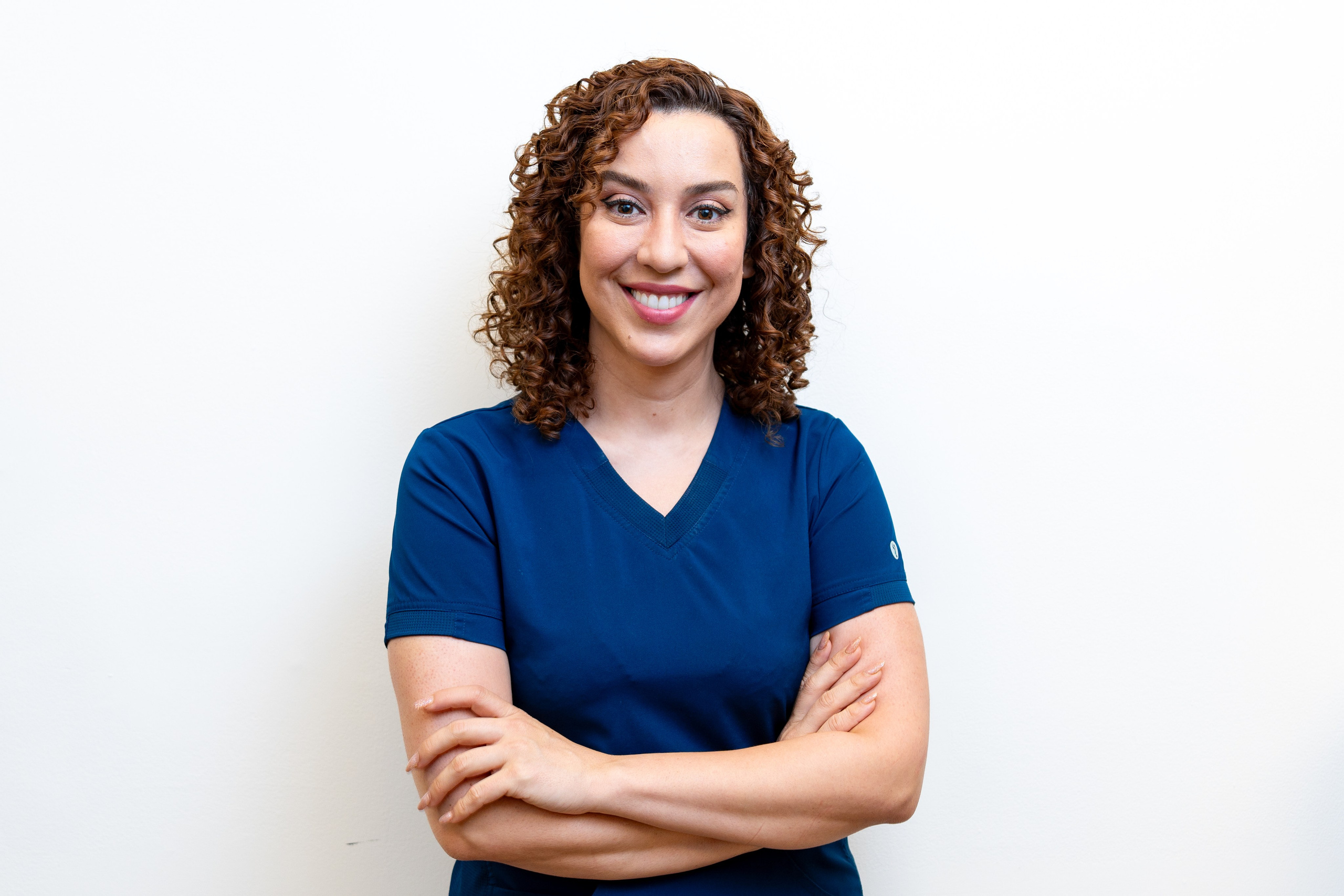 Portrait of a smiling female medical professional with curly hair, wearing navy blue scrubs, standing against a white background.
