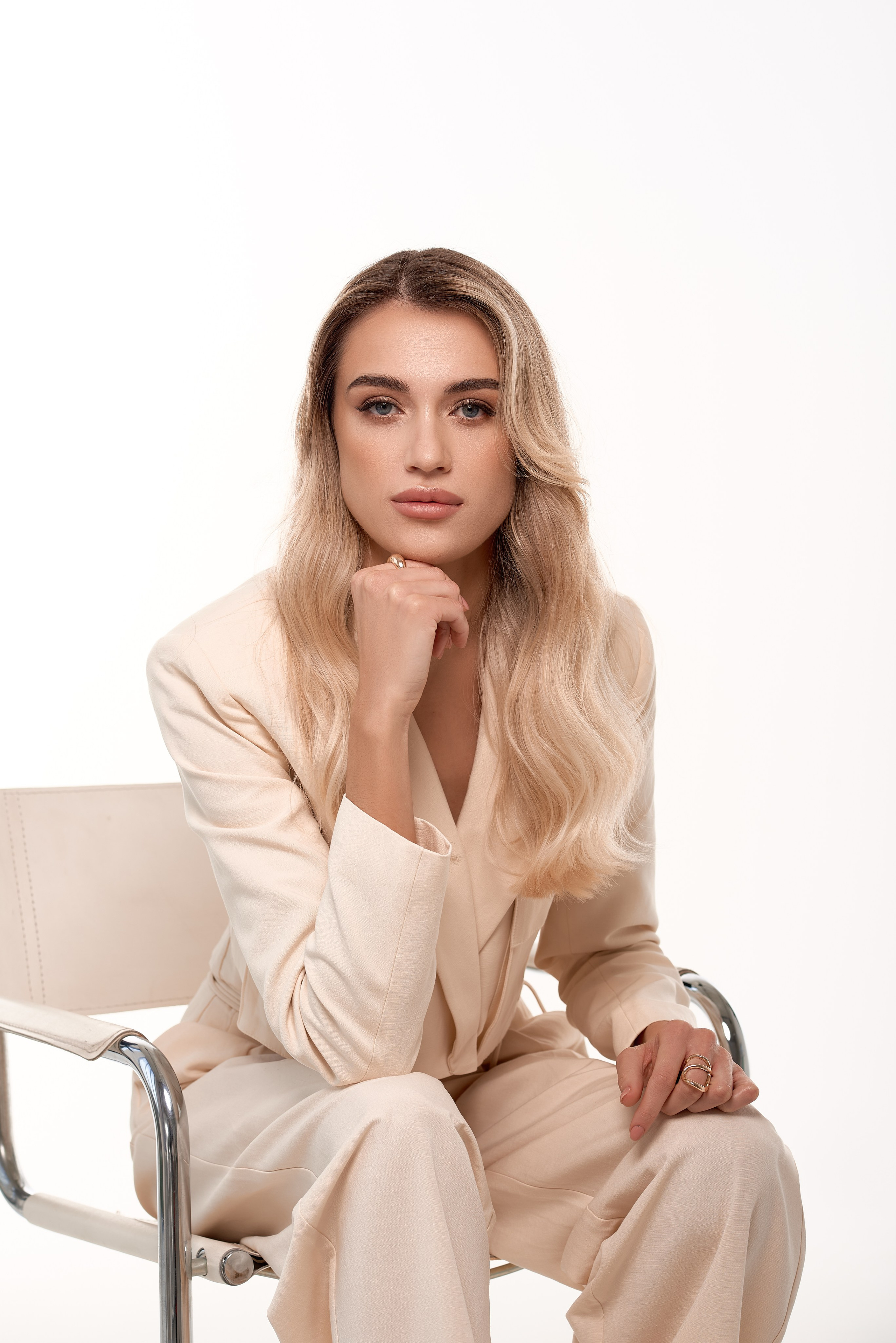 Жінка у кремовому брючному костюмі сидить на металевому стільці, випромінюючи впевненість та елегантність. Світлий фон та м'яке освітлення створюють професійний портретний кадр.