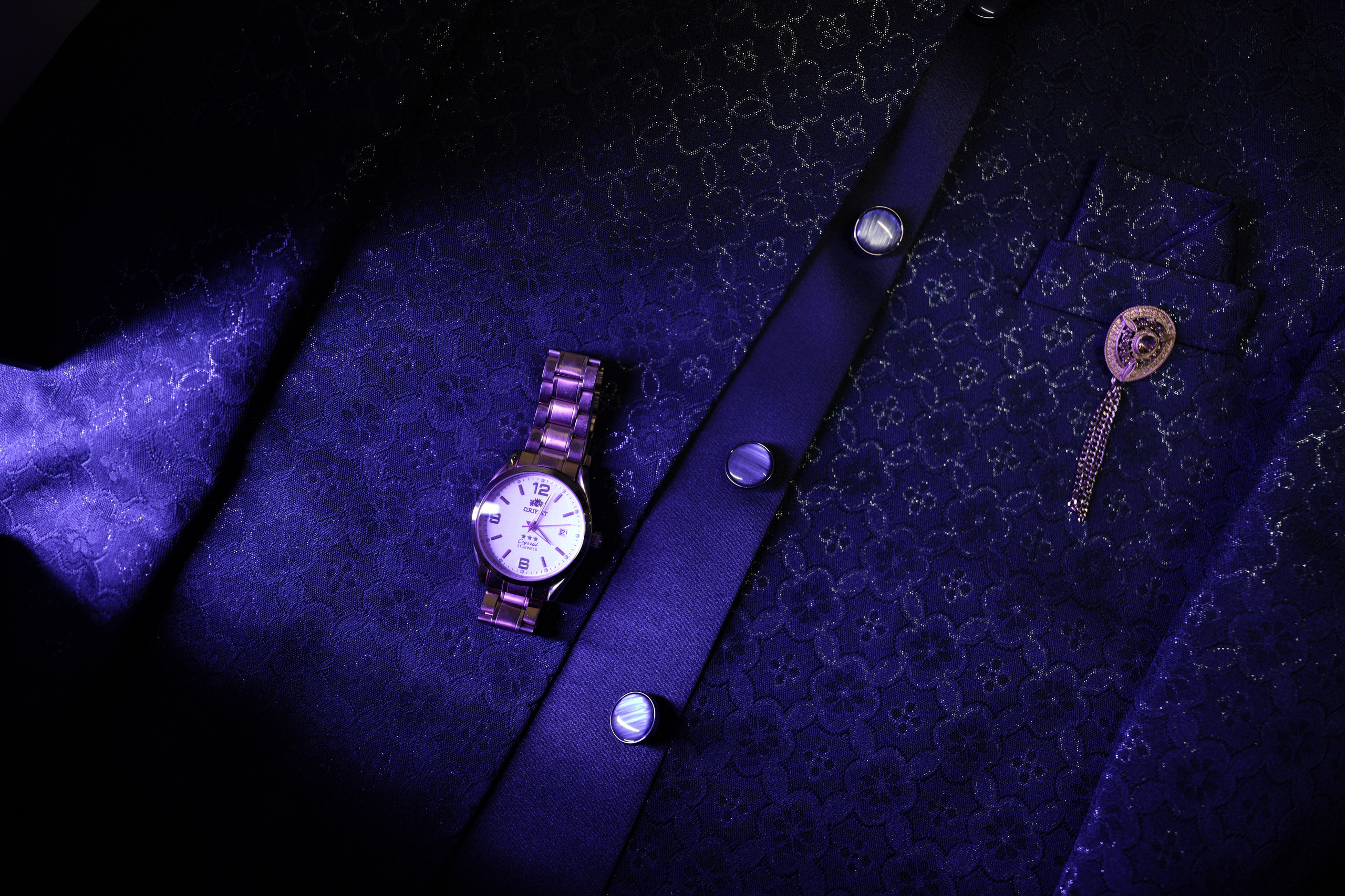 close-up product photo of a gold-tone wrist watch lying on a dark blue men's suit jacket with a golden lapel pin in Malleshwaram, Bengaluru