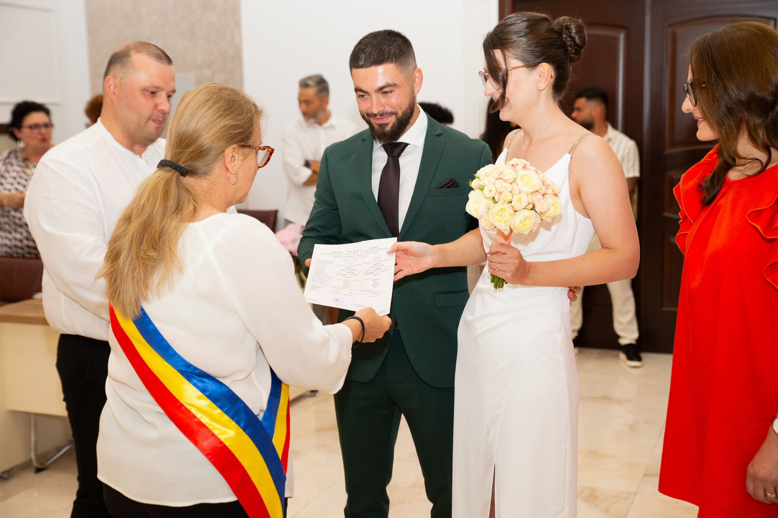 Bride in white dress and groom in dark green suit receiving marriage certificate from official with tricolor sash.