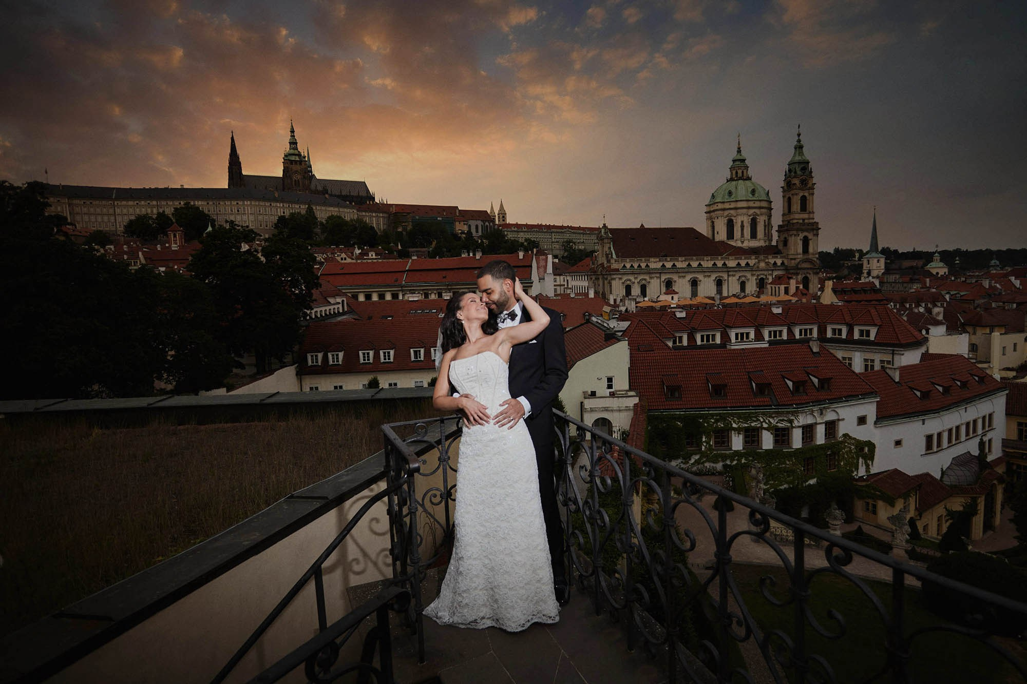 The Smiling Bride Leslie Caresses The Back Of Anthony's Head As She Leans Into His Arms Above The Vrtba Garden As The Sun Sets Behind Prague Castle.