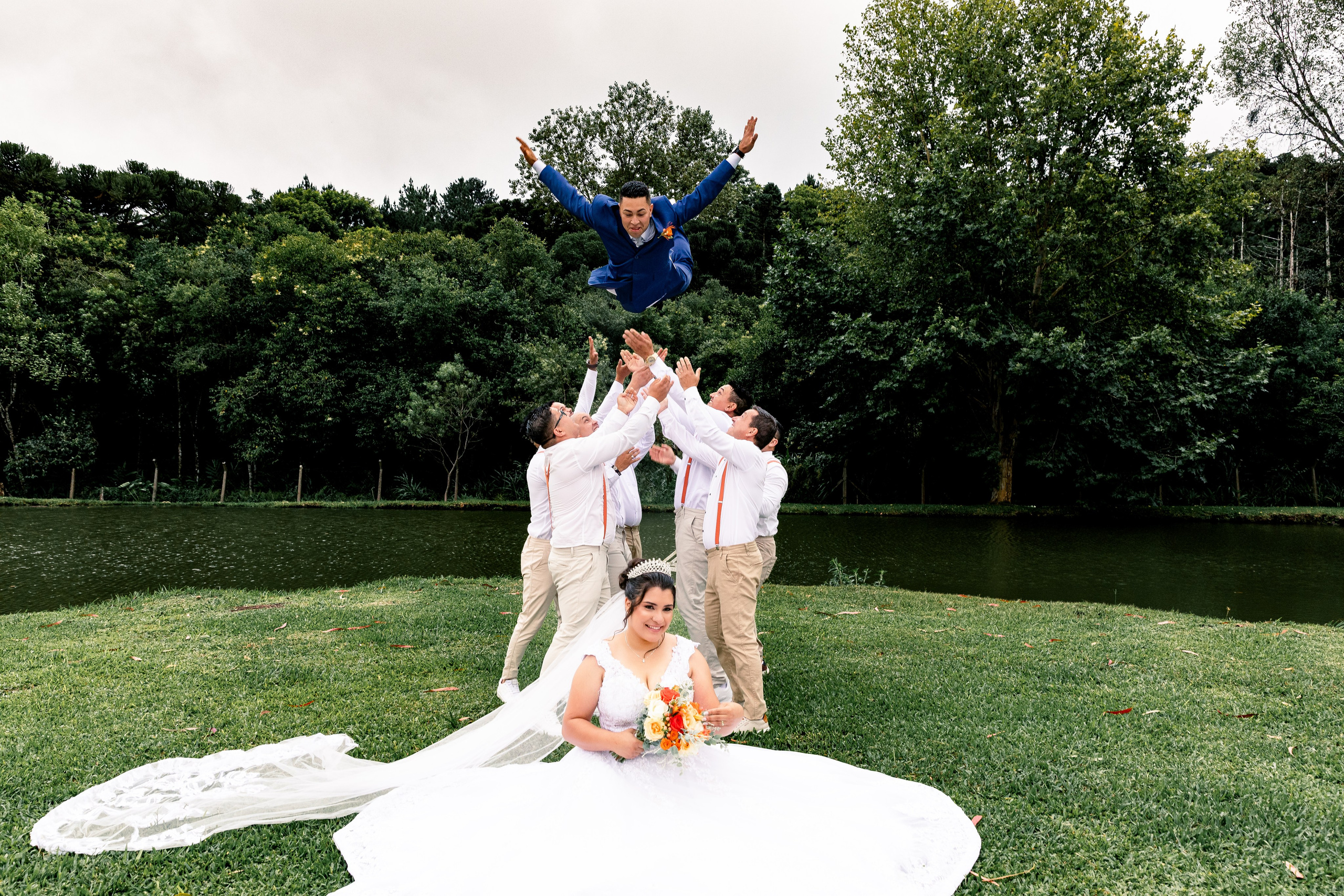 Noiva sorrindo com vestido esvoaçante enquanto o noivo pula em comemoração ao fundo com padrinhos.