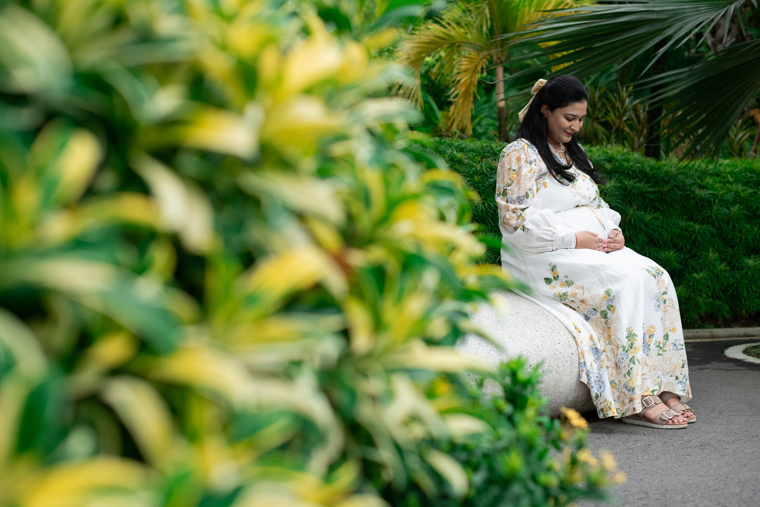 Pregnant woman in floral maxi dress sitting on a stone bench, surrounded by lush tropical greenery in Singapore botanical garden, gently holding her baby bump and smiling down.