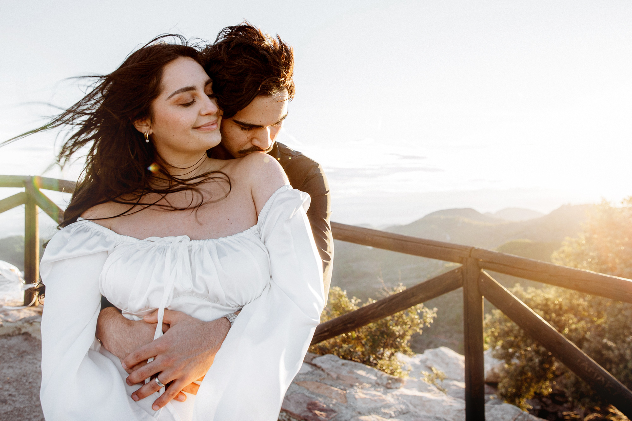 Engagement photoshoot in València, Spain, featuring a romantic couple embracing outdoors during golden hour, with soft natural light, gentle wind, and scenic hills in the background — an intimate and emotional engagement and wedding love story image ideal for engagement photography, pre-wedding photoshoots, romantic couple portraits, and professional wedding photography in València and across Spain.