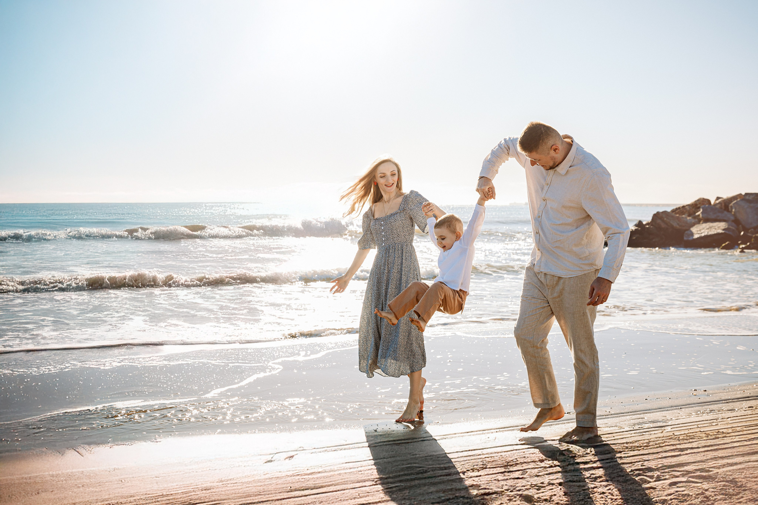 Alegre retrato familiar en una playa soleada de Valencia, España — padres balanceando a su hijo entre ellos junto al mar, capturando felicidad despreocupada y conexión junto al agua.