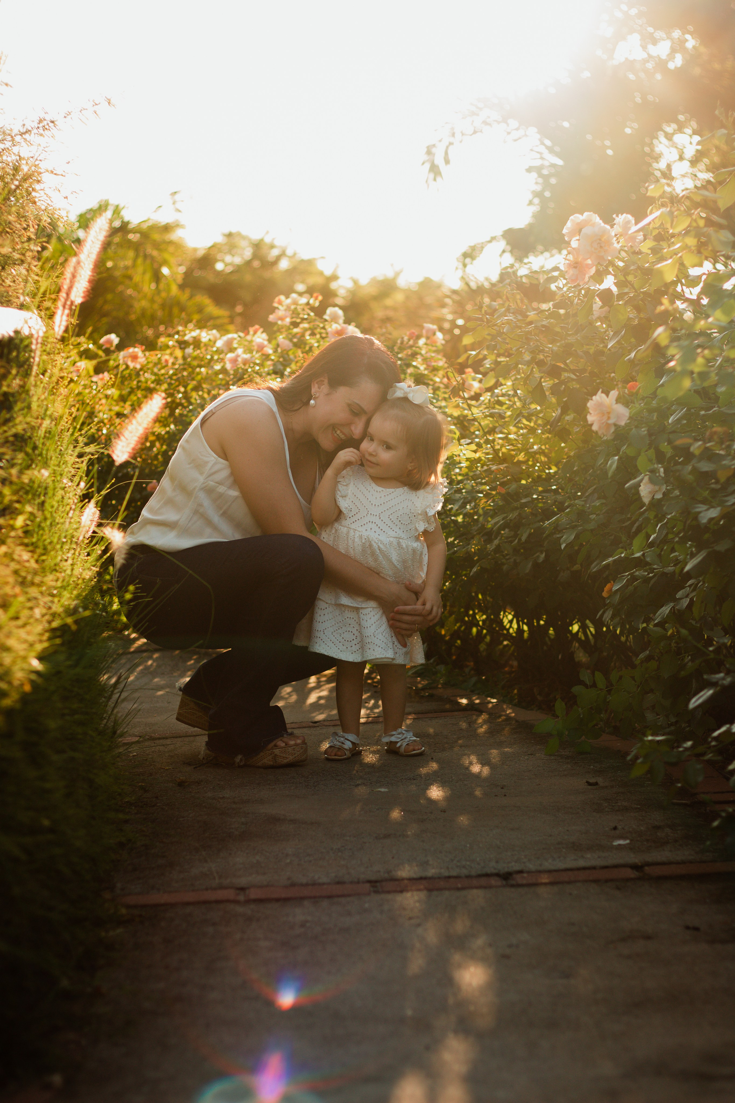 Mãe e filha com roupas brancas se abraçando no campo de rosas brancas em Holambra, durante ensaio ao entardecer com luz alaranjada.