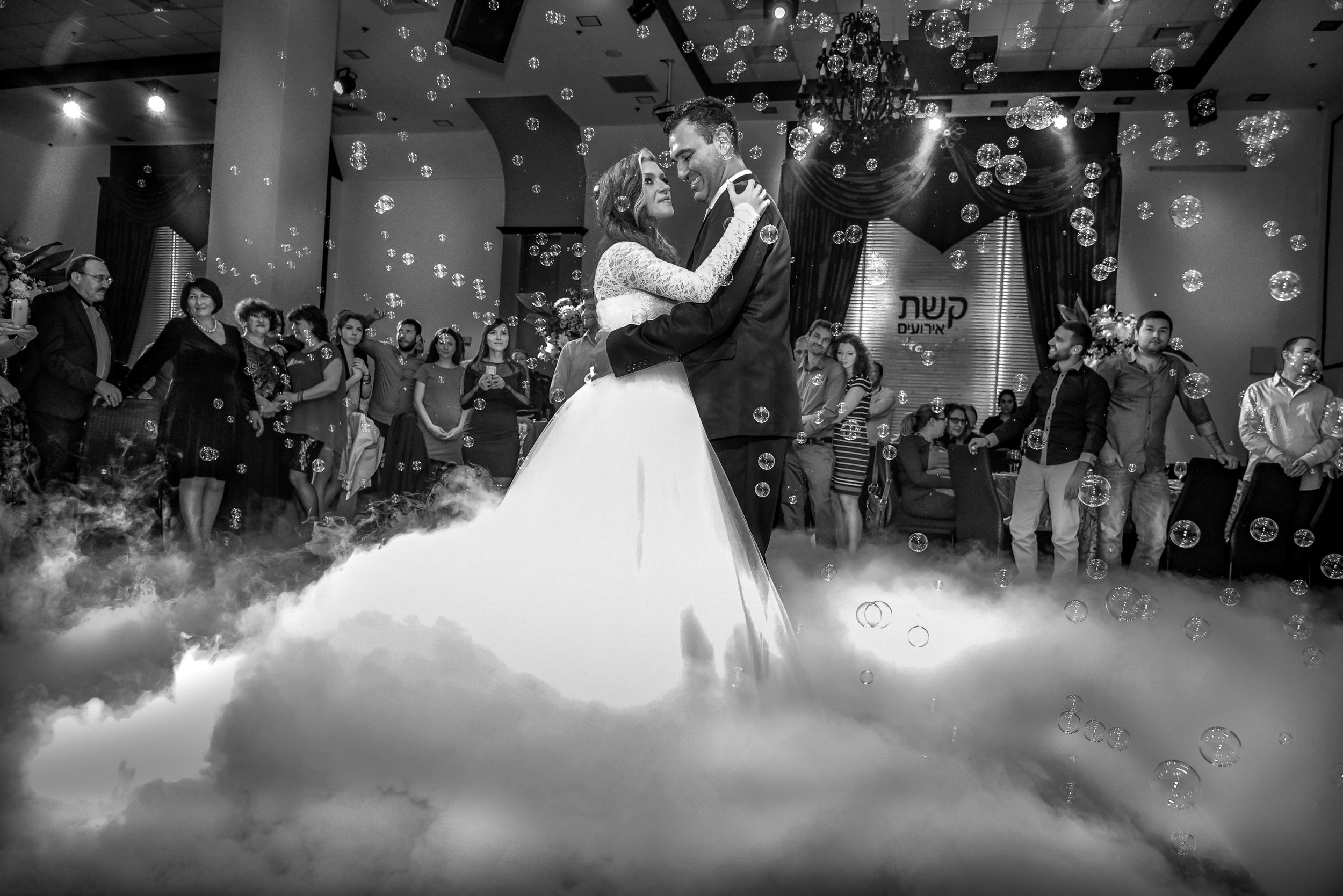 A black-and-white image of a couple's wedding dance, with the bride and groom embracing at the center of the frame, surrounded by guests and effects of artificial fog rising from the floor, and soap bubbles floating in the air.