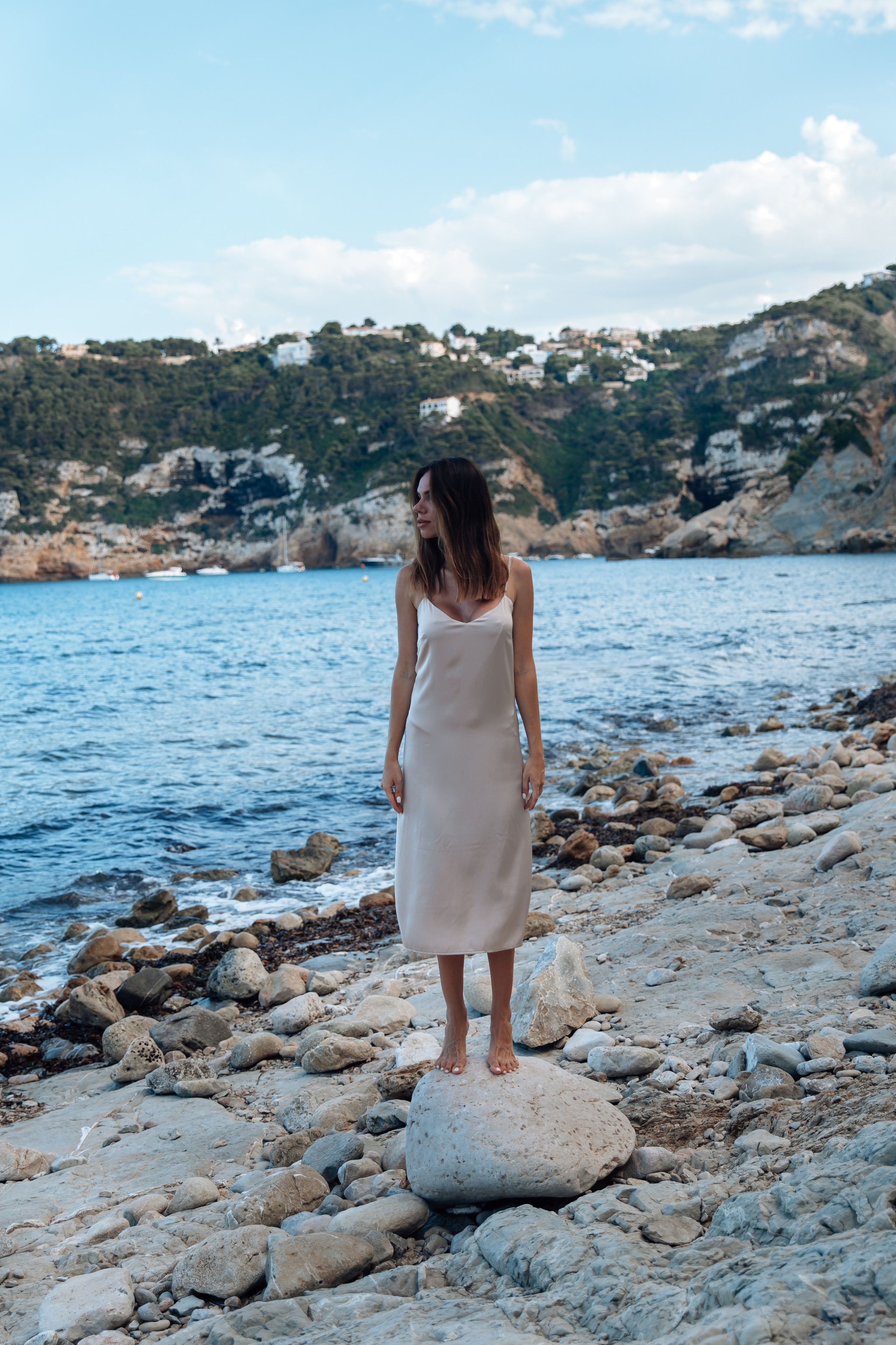 Женская фотосессия в Валенсии, Испания, с умиротворённым прибрежным портретом женщины, стоящей босиком на камнях у моря в лёгком платье, окружённой Средиземным морем и природным ландшафтом — идеально для женской портретной фотографии, лайфстайл-съёмок и профессиональных женских фотосессий в Валенсии и по всей Испании.