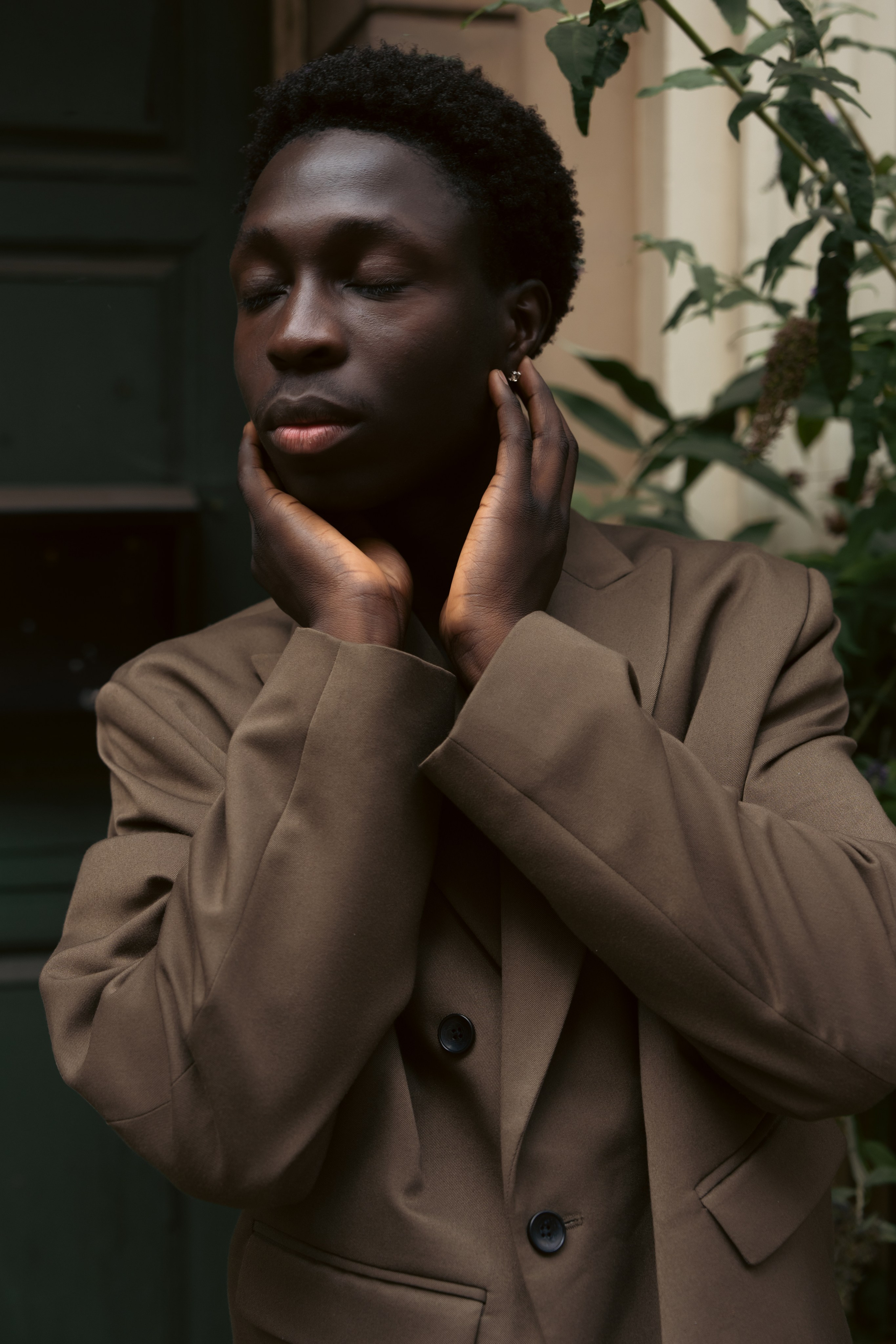 Editorial portrait of a male model in a brown suit surrounded by greenery