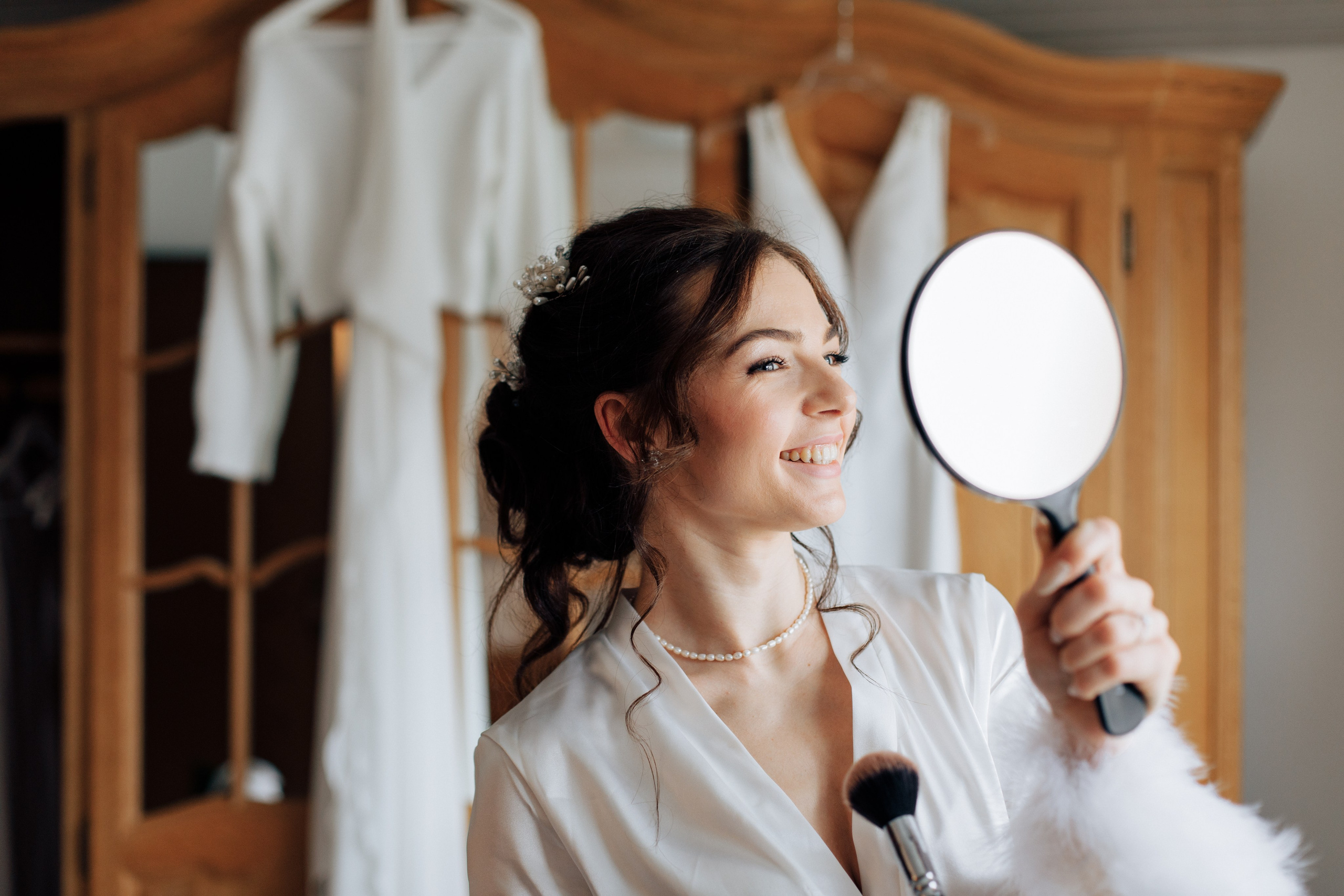 Die Braut blickt lächelnd in einen Handspiegel, während sie einen Make-up-Pinsel hält – im Hintergrund hängt ihr Hochzeitskleid.
