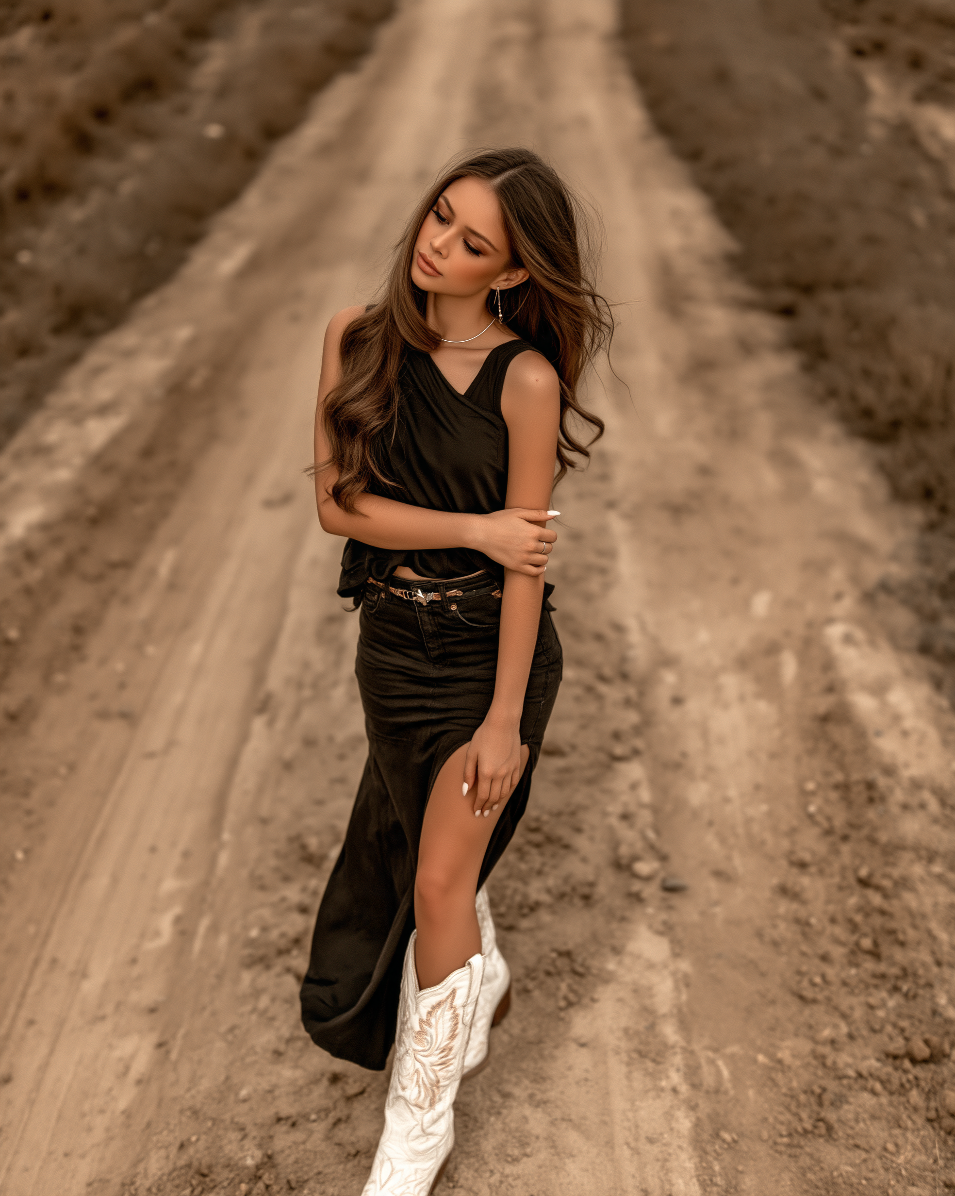 A moody, outdoor editorial portrait of a young woman with long, wavy light brown hair blowing in the wind. She is standing on a dusty rural dirt road with a dark, overcast sky and blurred landscape behind her. She wears a black sleeveless wrap top with an asymmetrical hem, a black denim skirt with a high side slit, a brown leather belt, and striking white cowboy boots with elaborate tan stitching. Her arms are crossed with one hand resting near her thigh, and she looks slightly upward and to the side with a calm, confident expression. The color grading is desaturated and warm, giving the image a cinematic, weathered feel
