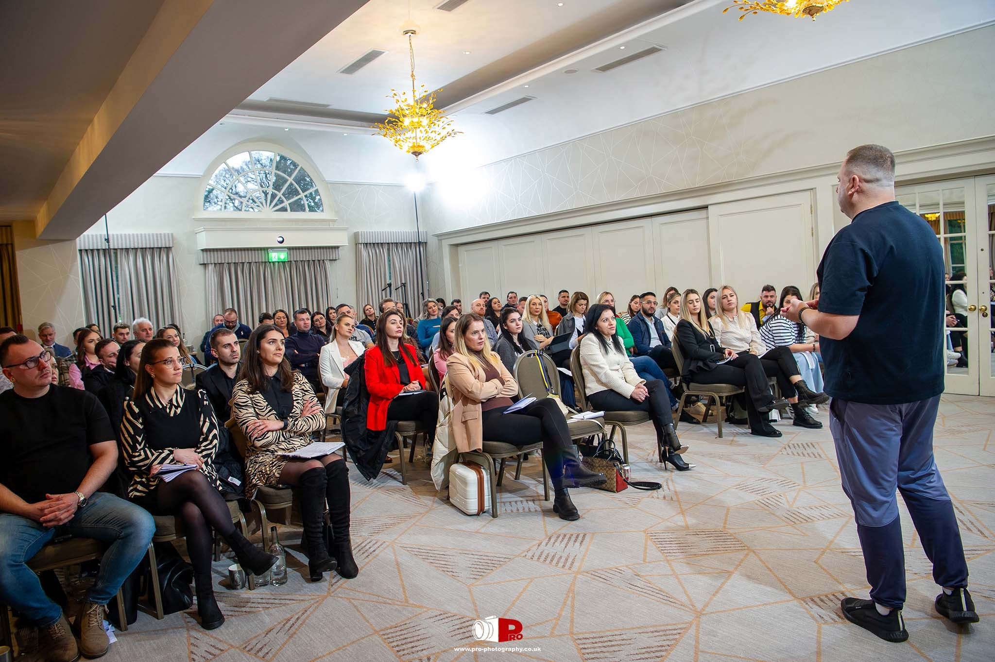 Audience members attentively listen to a speaker during a professional conference in a spacious hall with elegant decor.