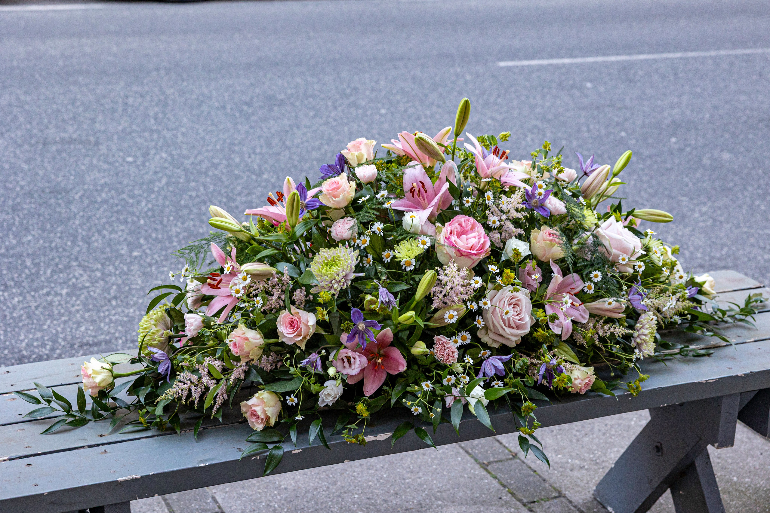 Denne kistepynt kombinerer liljer, roser og markinspirerede blomster i lyse nuancer. Akropolis Blomster skaber personlige begravelsesdekorationer med fokus på kvalitet, omsorg og æstetik.