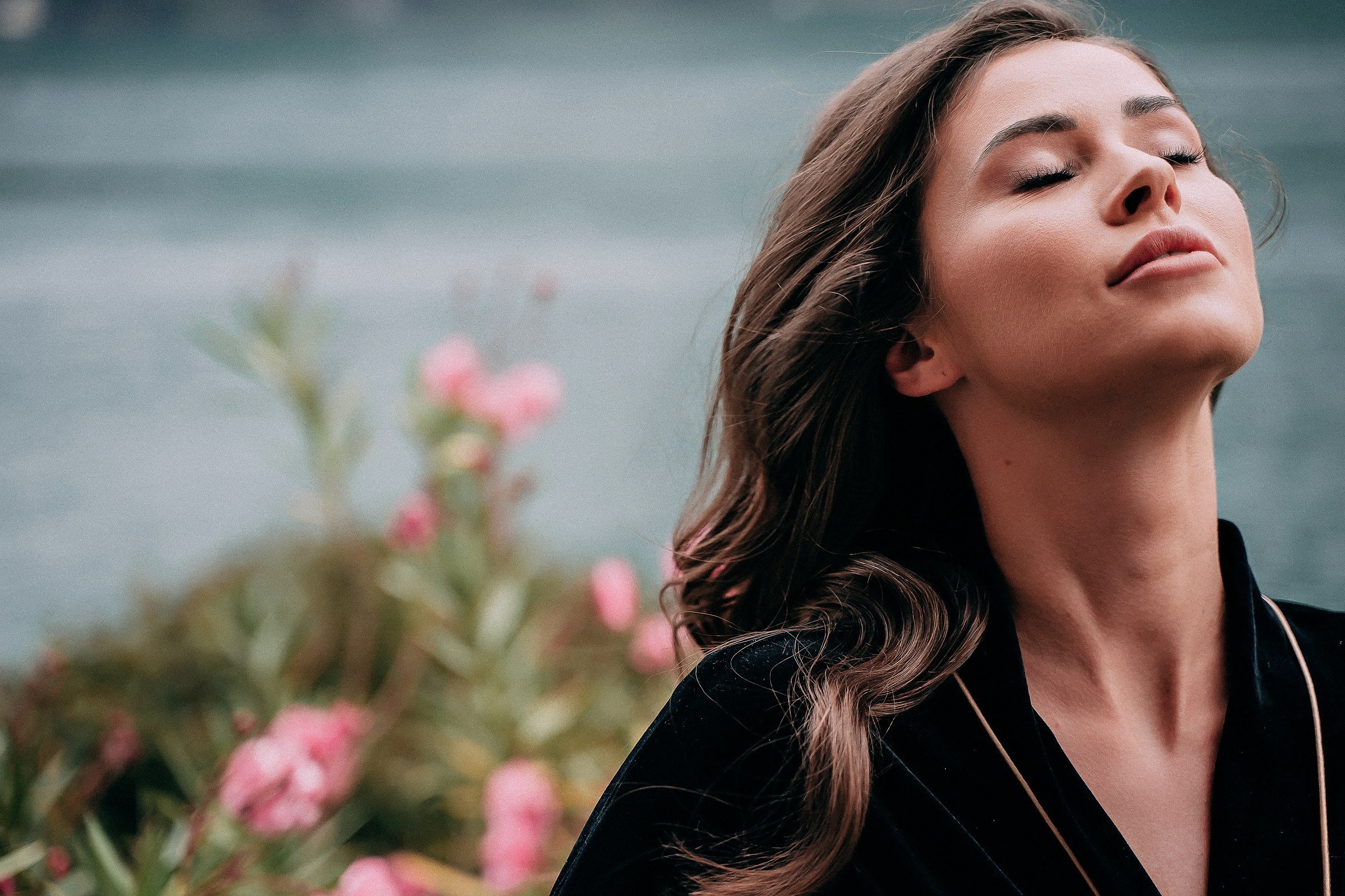 Close-up of a bride with long, wavy hair wearing a dark garment with light trim, standing near a lake. Pink flowers and lush greenery frame the serene background of water and soft natural light.