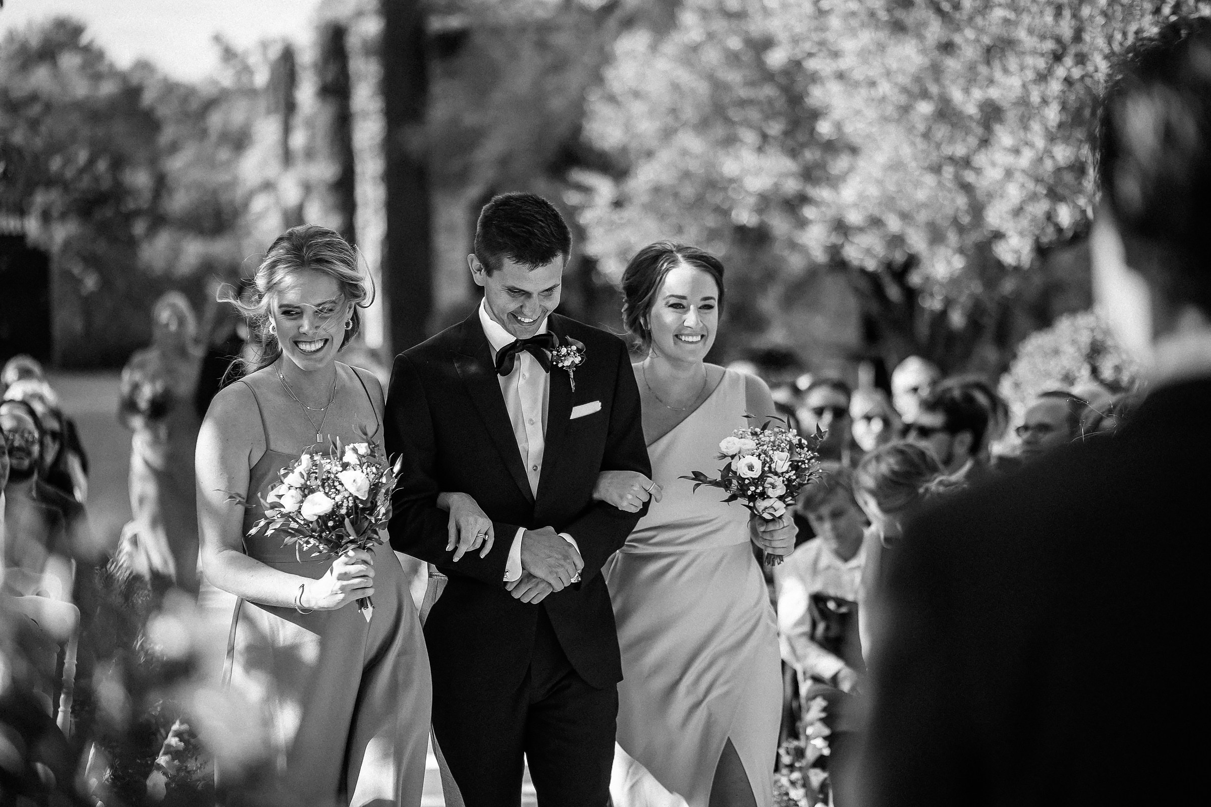 A black-and-white capture of the trio walking down the aisle, surrounded by guests and sunshine at the outdoor wedding ceremony. 