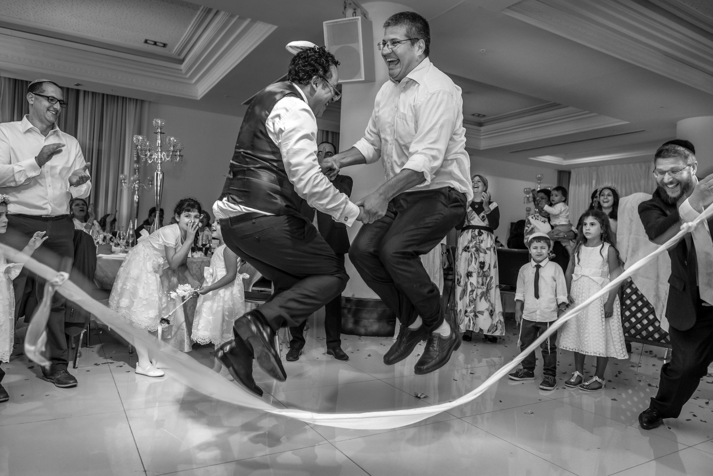 An electrifying moment of celebration—two men leap joyfully over a ribbon, their laughter echoing through the room. The energy is contagious, drawing smiles from guests of all ages.