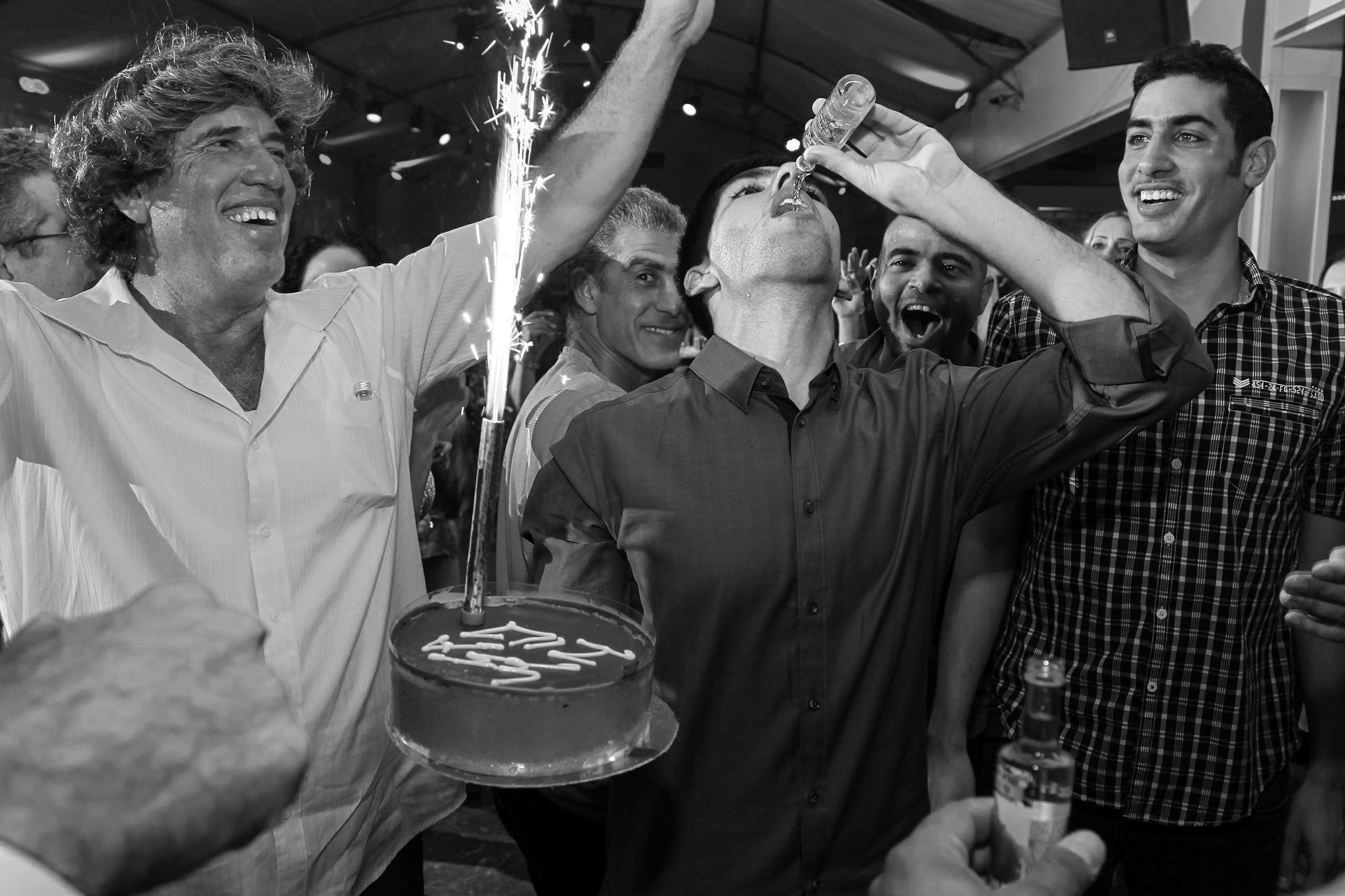 A dynamic and joyful celebration captured in black and white: A man raises a cake with a lit sparkler while another enthusiastically takes a shot, surrounded by cheering friends. The energy, laughter, and spontaneous emotions make this moment feel alive and unfiltered.