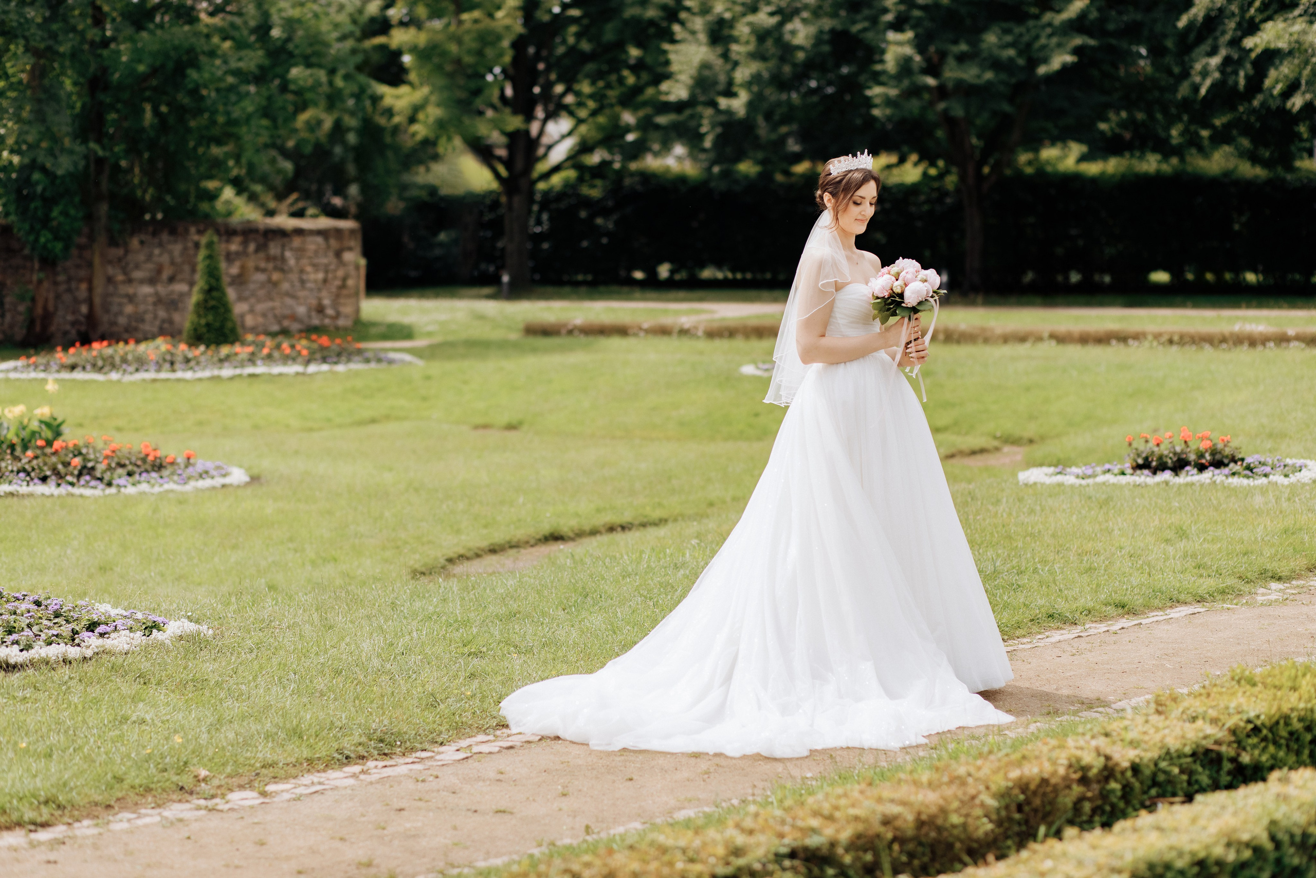 Die Braut steht in einem schönen Garten des Schlosses Heusenstamm, trägt ein elegantes Hochzeitskleid mit offenen Schultern