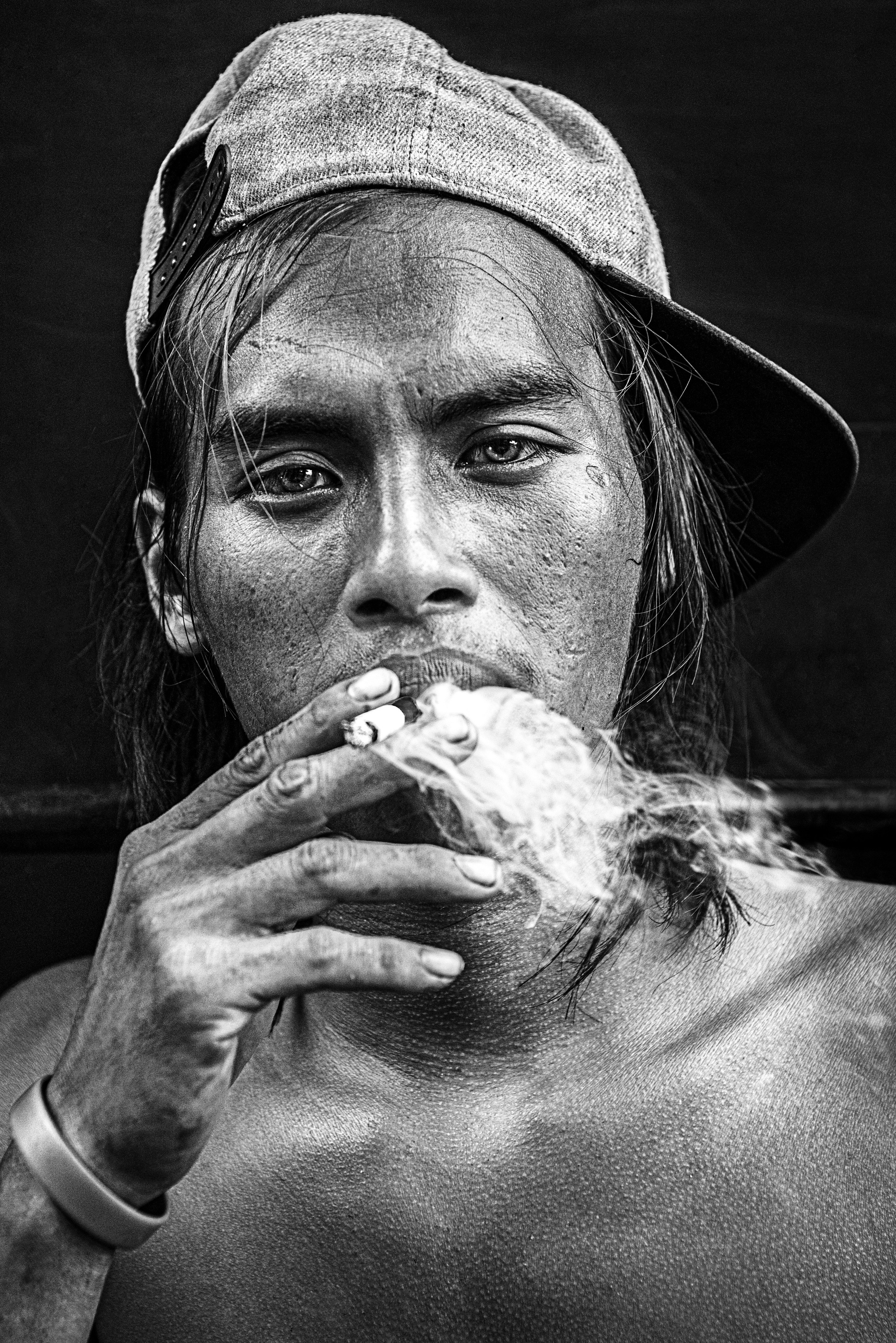 A gritty black-and-white portrait of a man smoking a cigarette, with smoke curling around his face. Wearing a backward cap and staring directly at the camera, his weathered features and intense gaze convey raw emotion and authenticity. This image showcases the photographer’s ability to capture candid and powerful portraits, perfect for clients looking for bold and expressive storytelling in editorial, documentary, or branding work.