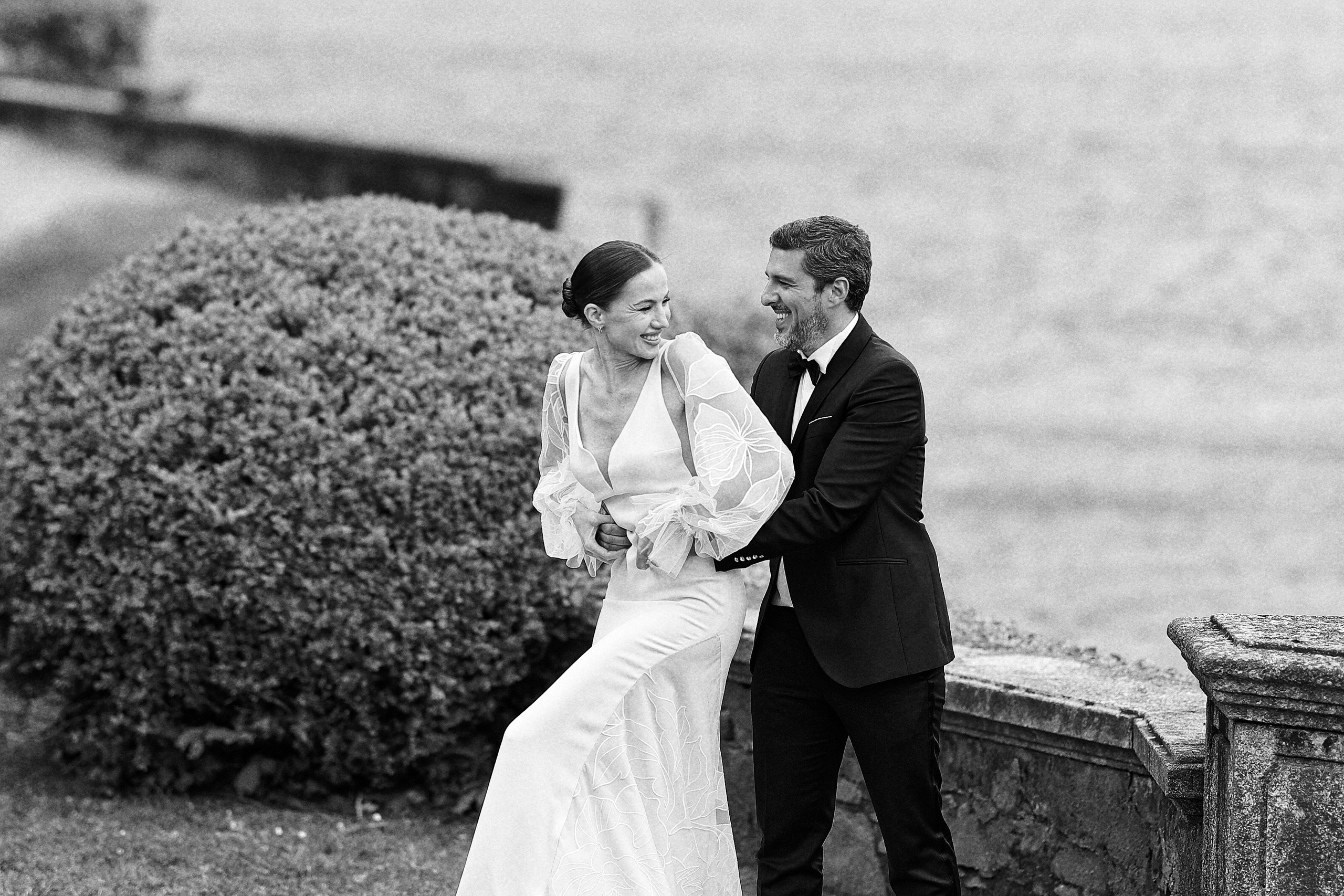 "Black and white shot of a bride and groom laughing and embracing along a lakefront promenade."