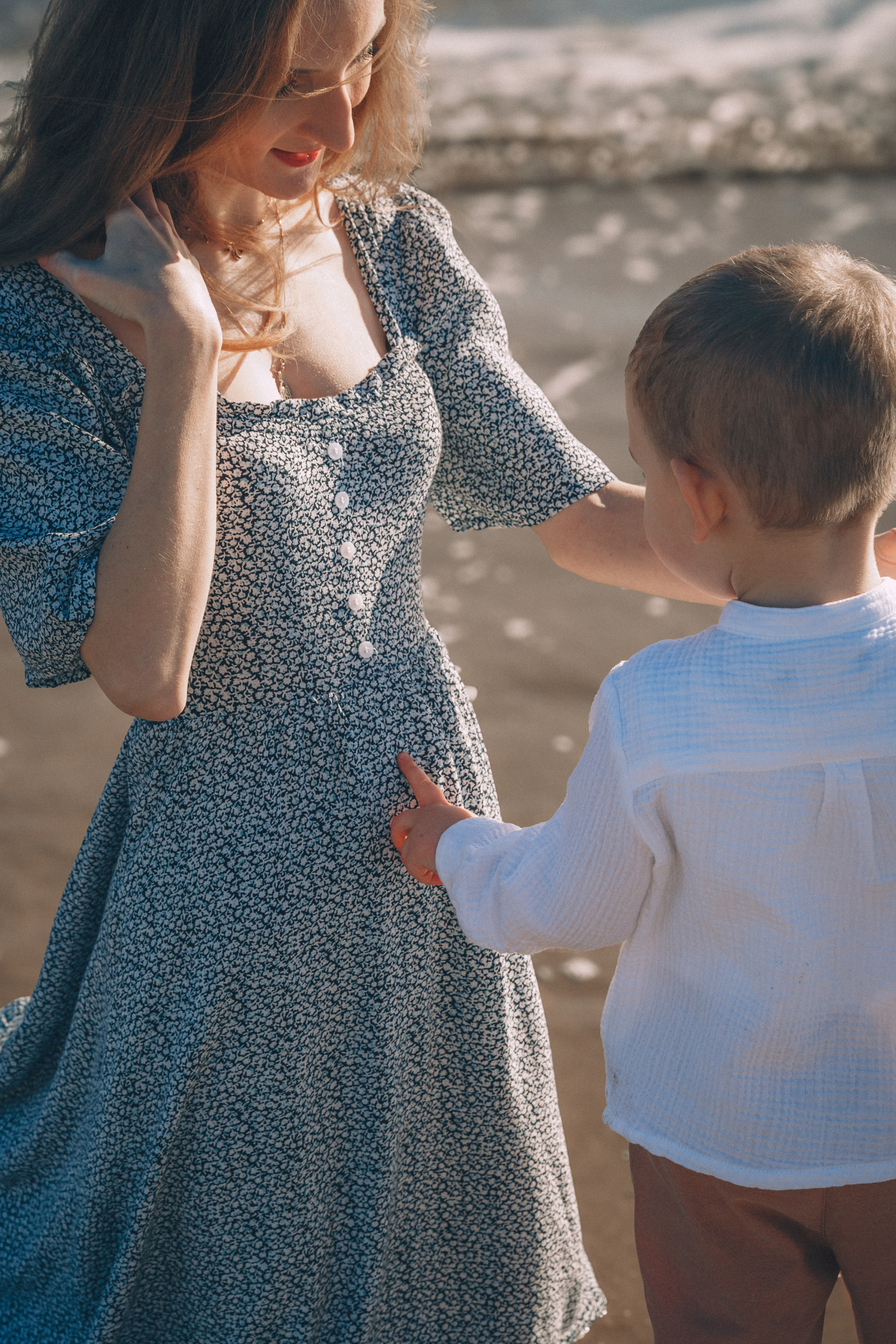 Sesión de fotos de embarazo en València, España, capturando un tierno momento familiar en la playa mientras un niño pequeño toca suavemente la barriga de su madre embarazada, fotografiada con luz solar cálida y natural, tonos mediterráneos suaves, reflejos del mar y una fuerte conexión emocional — ideal para fotografía de embarazo, sesiones de maternity, sesiones familiares de embarazo, retratos lifestyle de embarazo y sesiones de embarazo en la playa en València y en toda España.