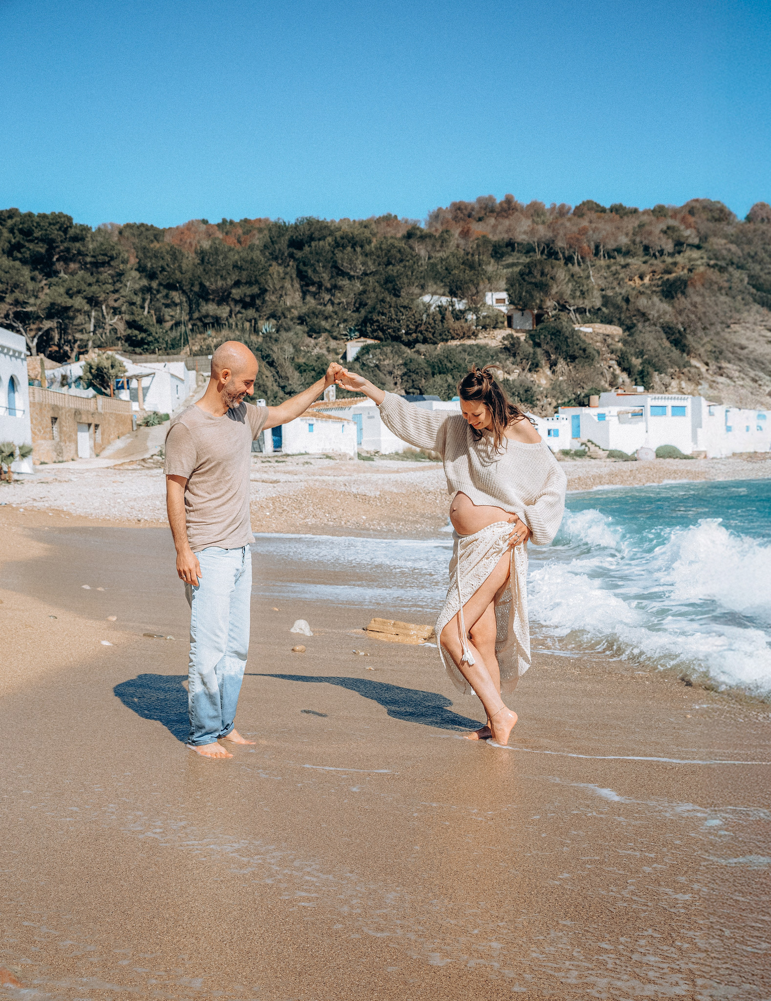 Foto alegre de embarazo capturada en la playa soleada de Valencia, España — pareja descalza bailando en la orilla, la futura mamá con suéter acogedor y pareo de encaje, sonriendo mientras las olas tocan sus pies.