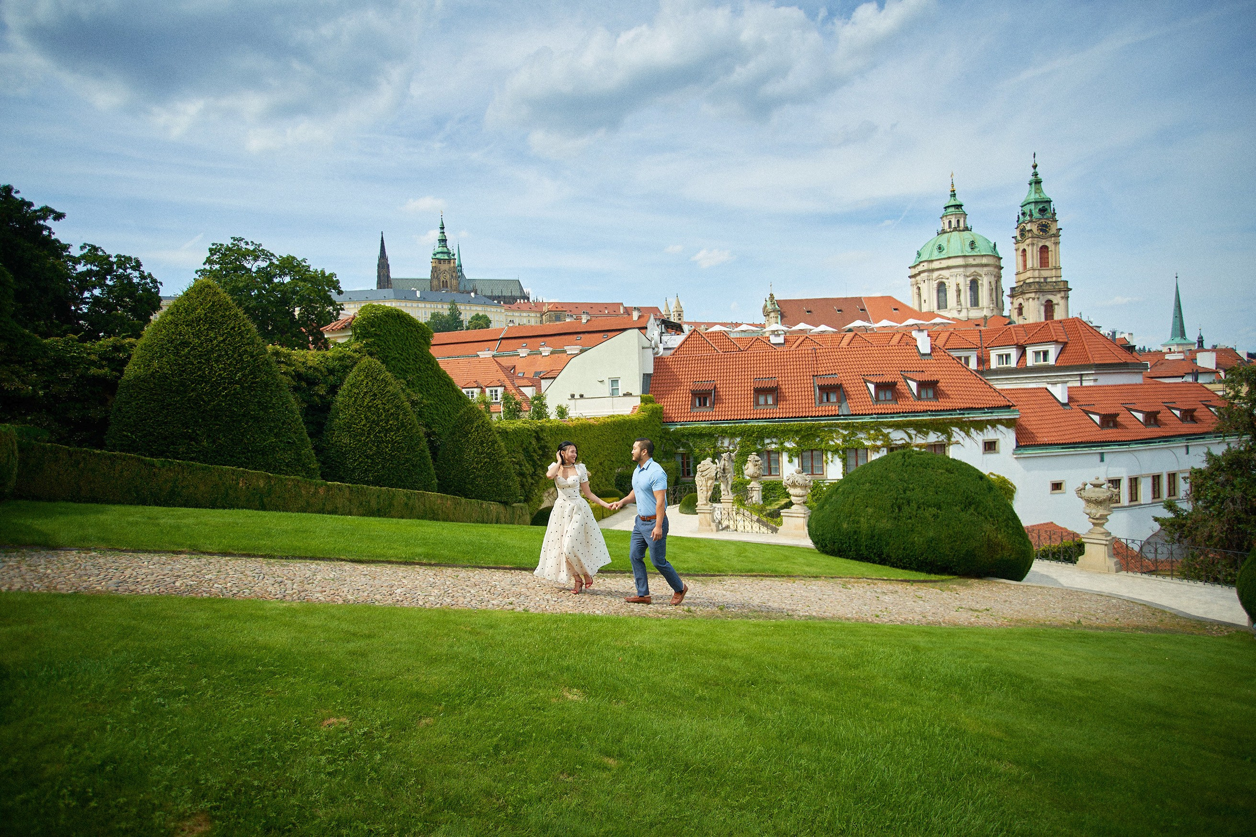 Wide view of engaged couple walking with Prague Castle and St. Nicholas Church dome in the background from Vrtba Garden.