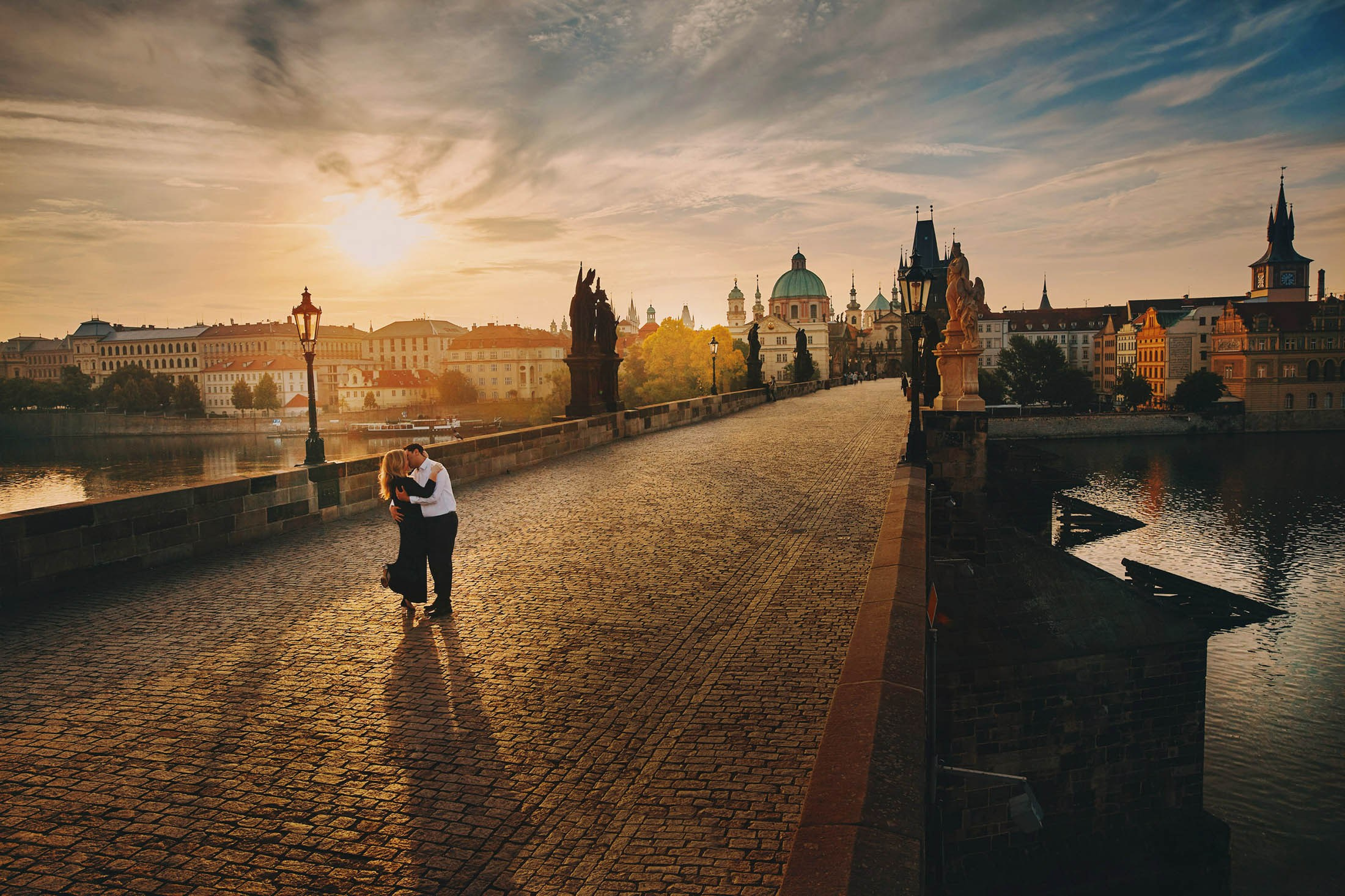 Couple sharing a romantic kiss on Charles Bridge with sunrise lighting over Prague