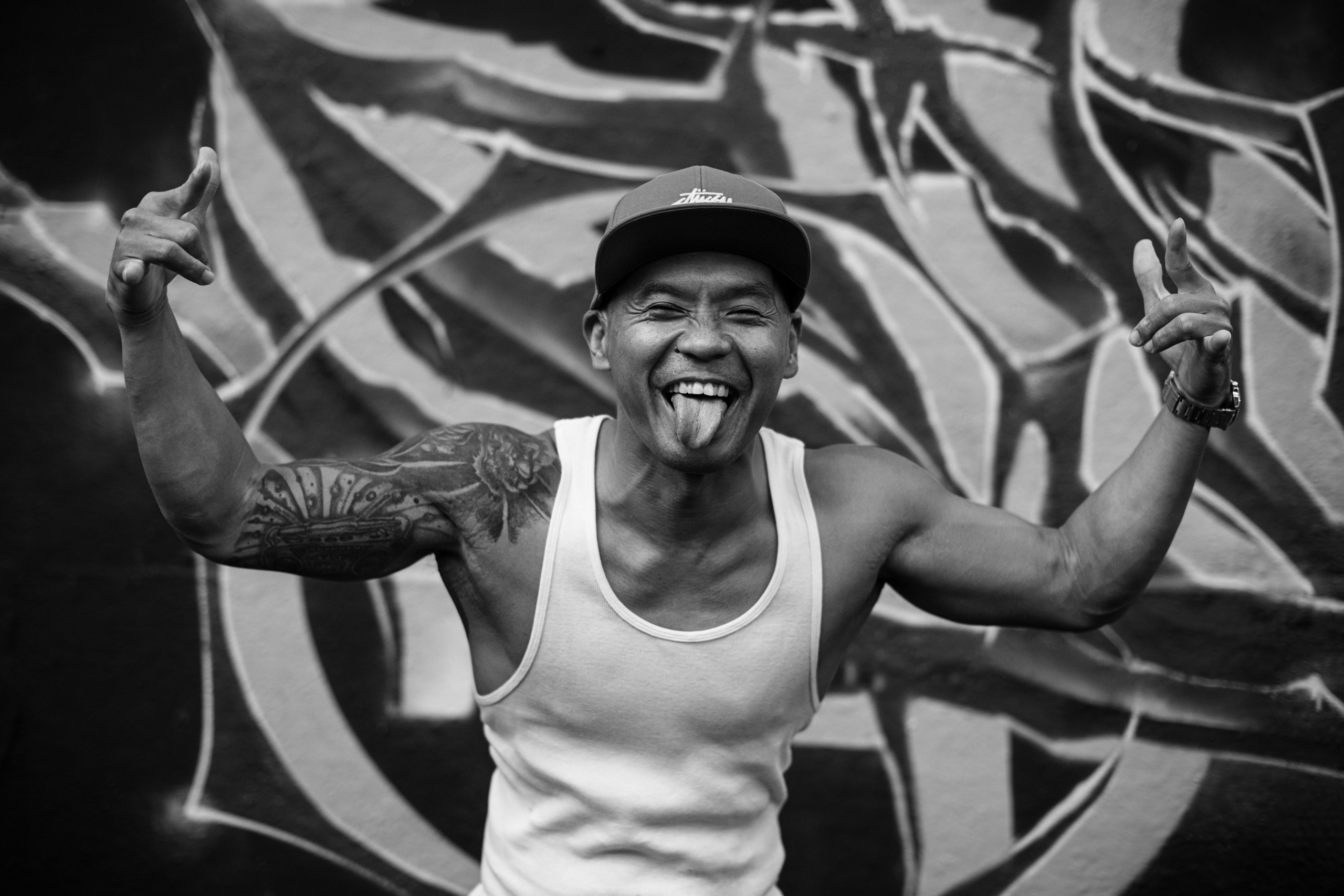Energetic black‑and‑white street portrait of a tattooed man posing playfully before a graffiti mural—captured by Singapore professional photographer Max&nbsp;Mirrorklezz; showcases dynamic, candid style available for bookings and personal branding sessions.