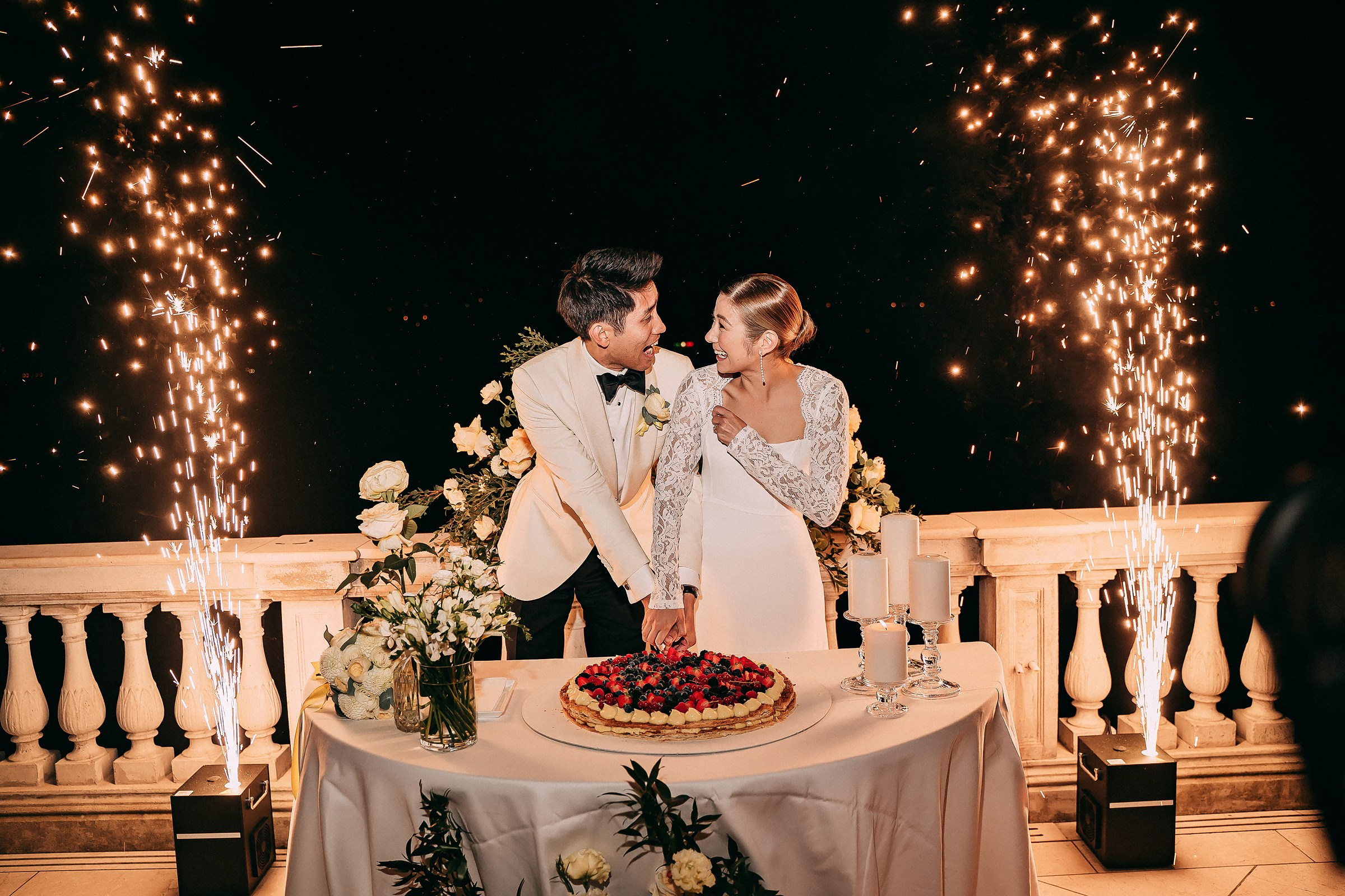 Bride and groom share a playful glance while cutting the wedding cake under a shower of sparklers.