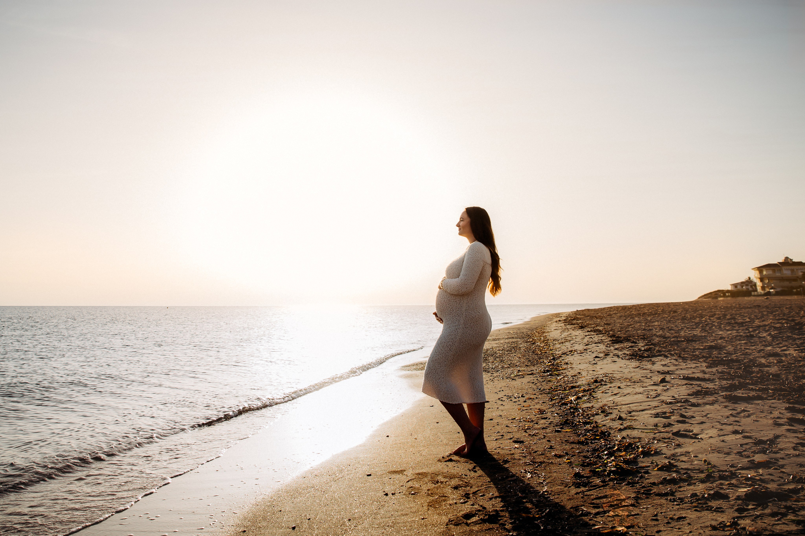 Serena sesión de maternidad en la playa de Barcelona, España — mujer embarazada con vestido ajustado contempla el mar al atardecer. Perfecta para quienes buscan fotografía de embarazo elegante e íntima en Barcelona y la costa española.