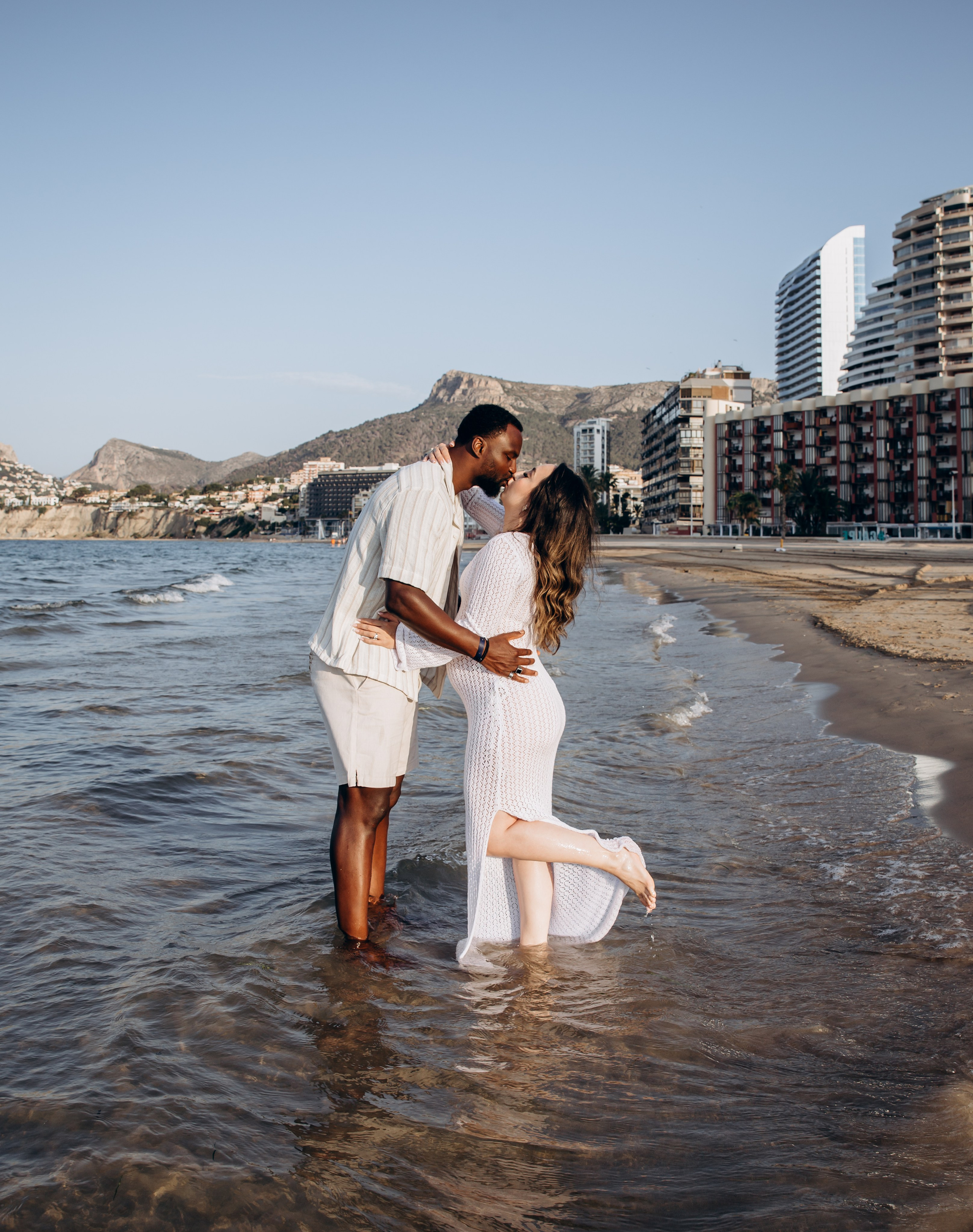 Sesión romántica de embarazo en Calpe, España — pareja compartiendo un beso en el mar con el horizonte de Calpe y las montañas al fondo. Una sesión soñada para quienes buscan fotografía de maternidad íntima y escénica en la Costa Blanca.