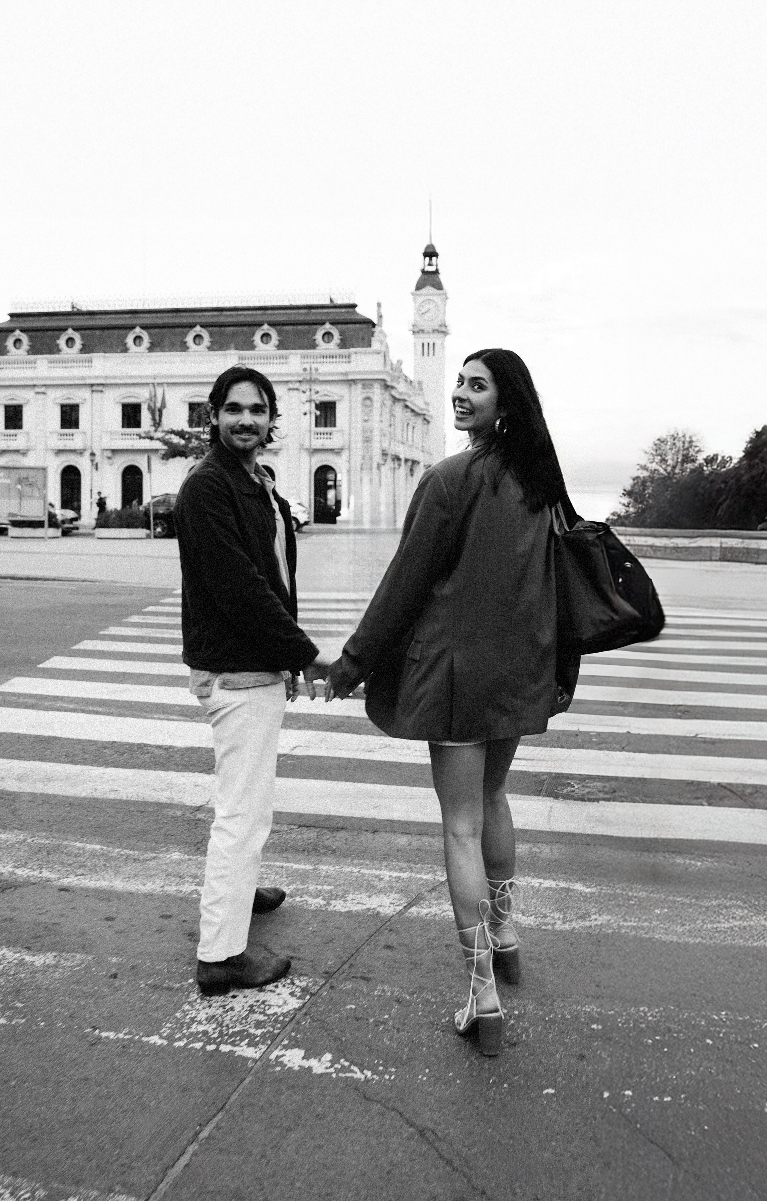 Romantic black and white couple photo taken during a love story photoshoot in Valencia, Spain — featuring a joyful moment as the couple holds hands crossing the street near the historic Clock Tower. Perfect for those searching for candid couple photography in Valencia or timeless love story photoshoots in Spain.
