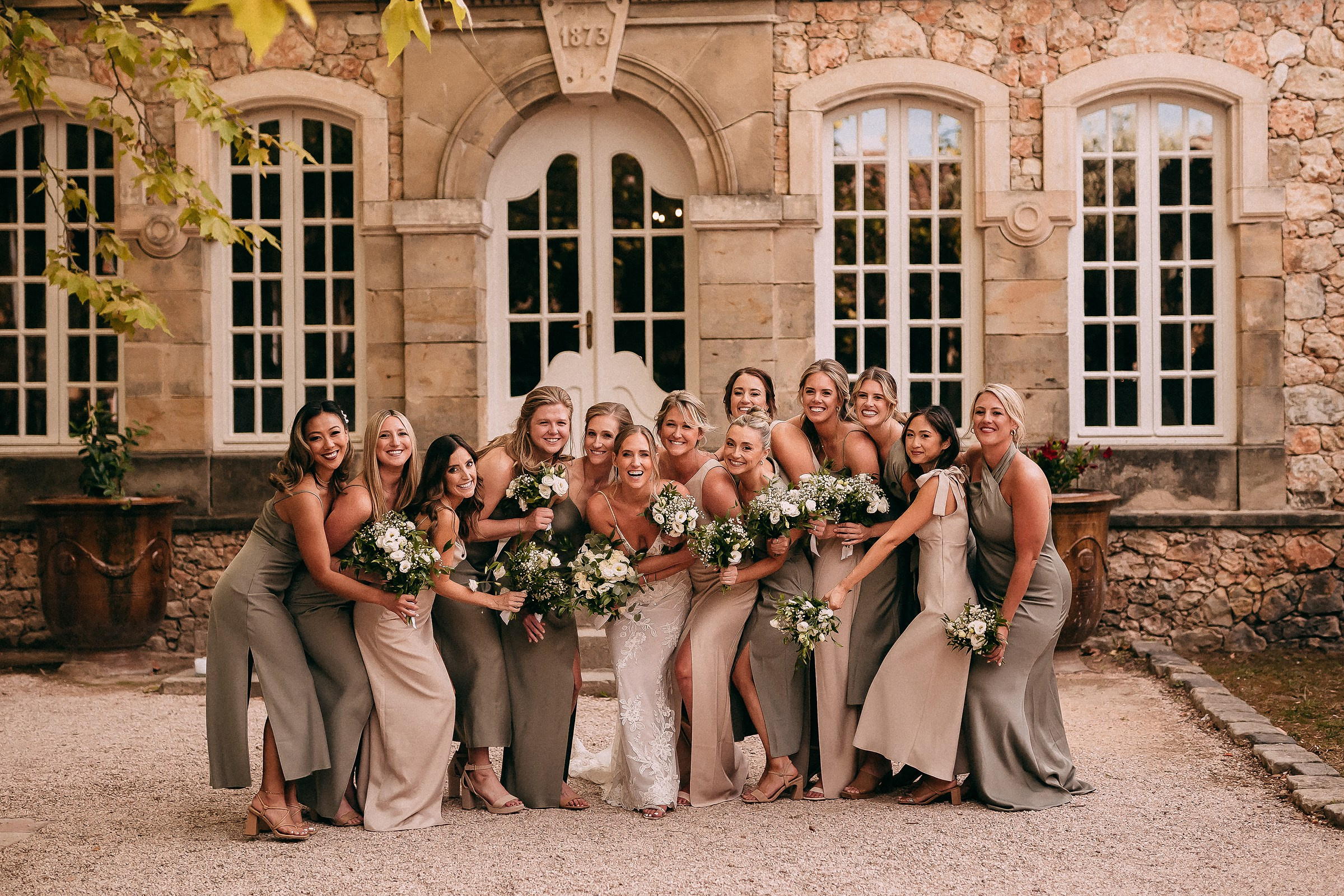 The bride and her bridesmaids gathered in front of a charming stone building, laughing and holding bouquets in varying shades of green and white.