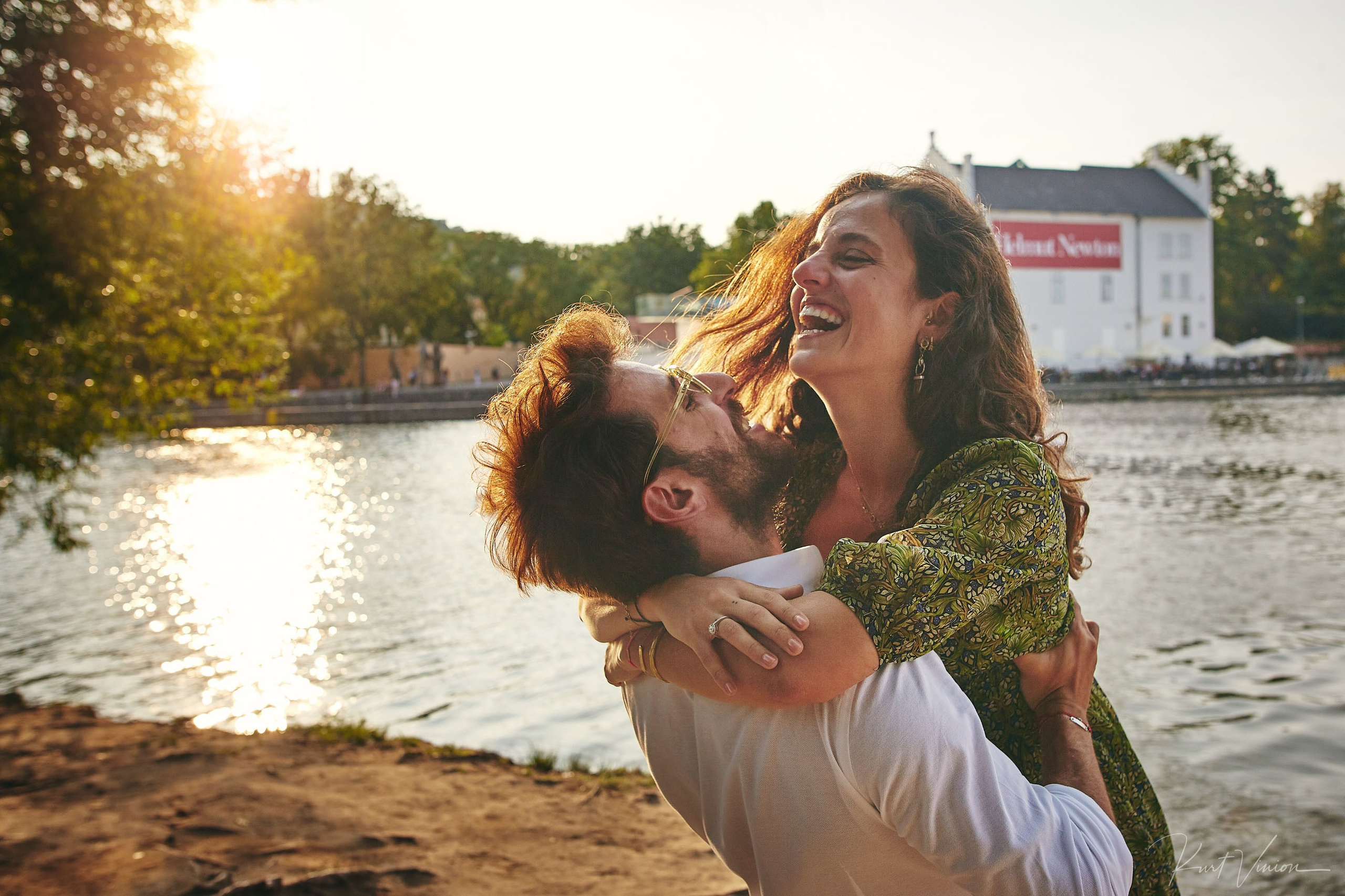 Man wearing sunglasses spins laughing fiancée in the sunlight by Kampa riverbank in Prague.
