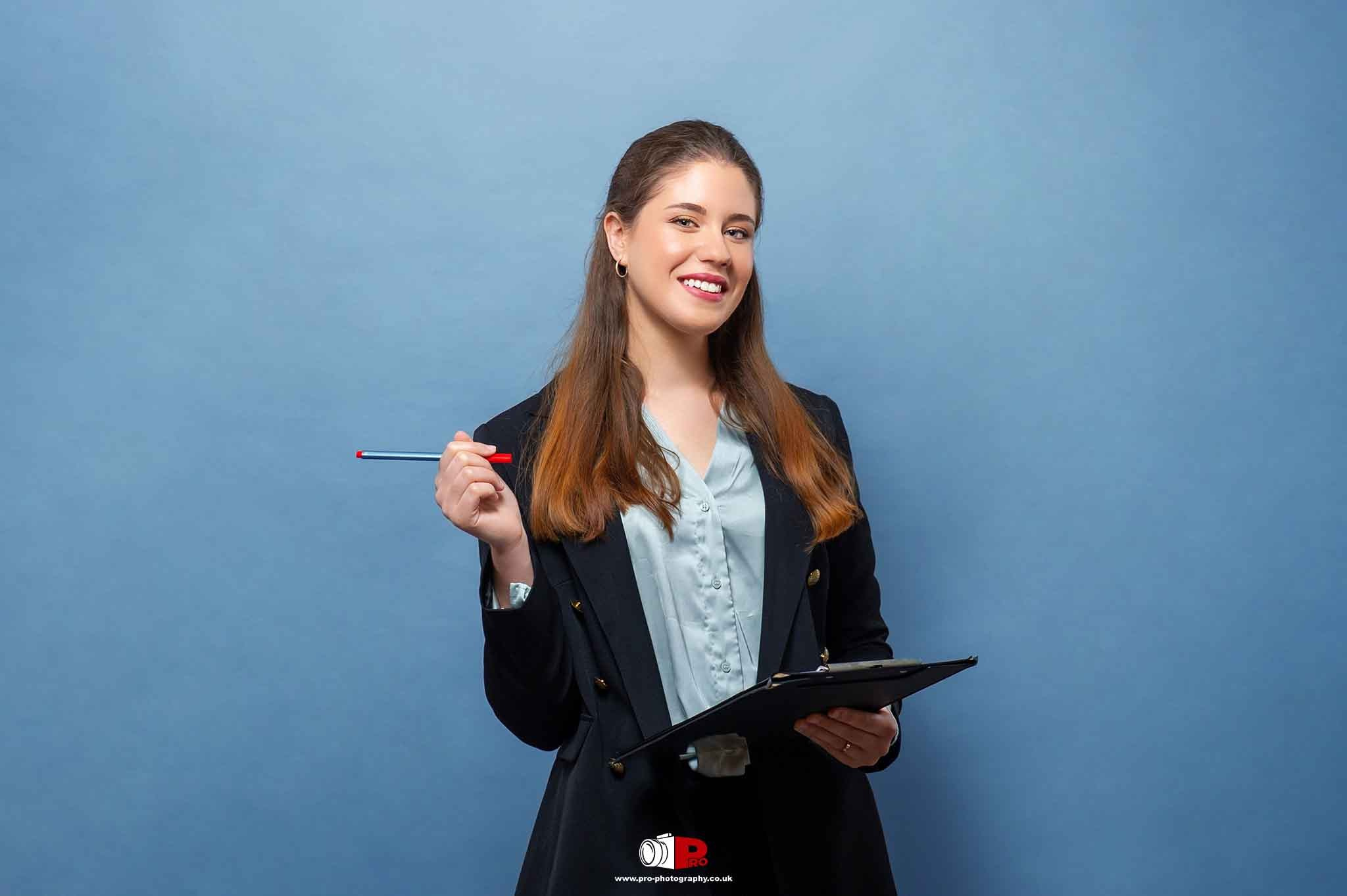 A confident young woman in a black blazer holding a clipboard and a red pen, posing against a light blue background.