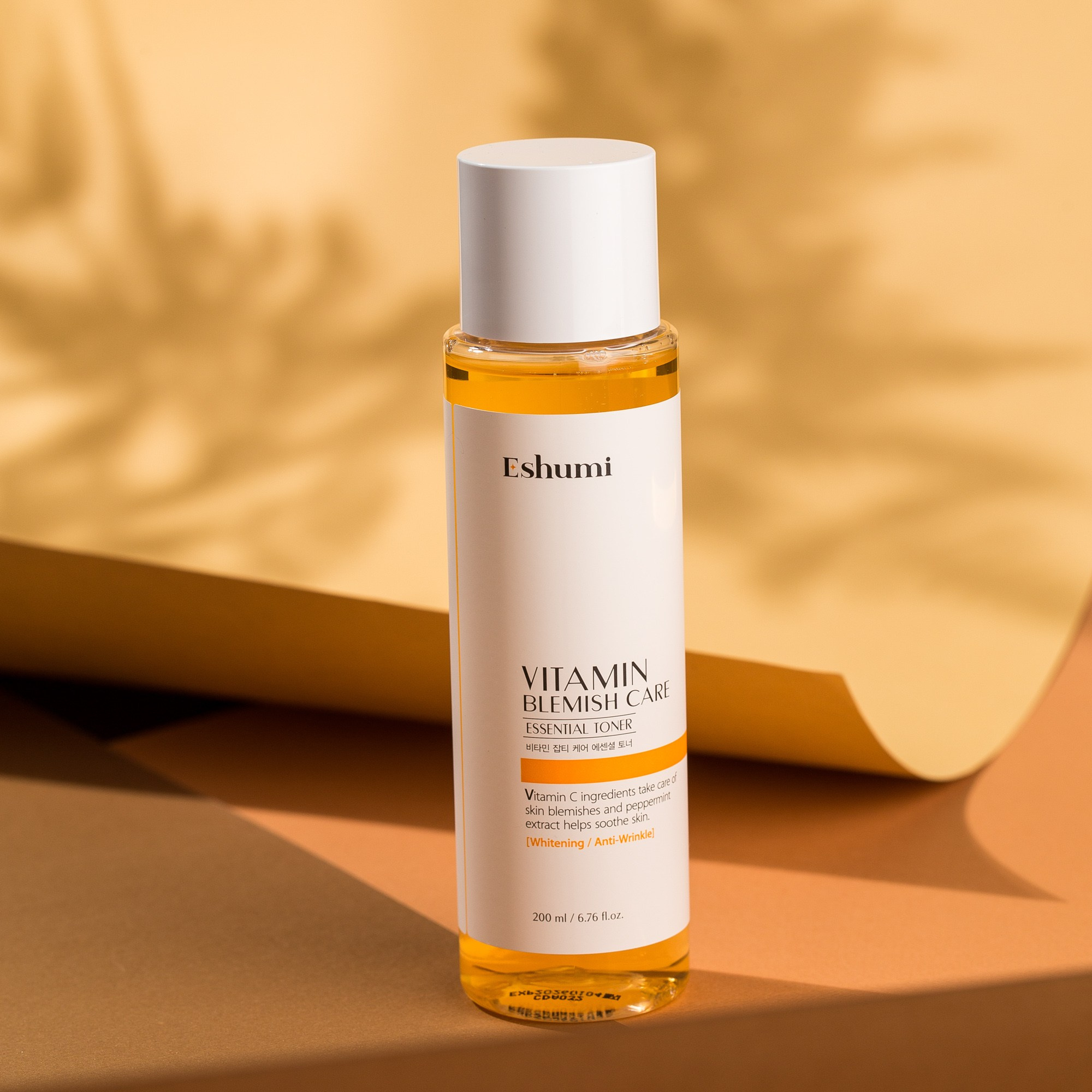 Eshumi Vitamin Blemish Care Essential Toner şüşəsi, sarı və narıncı fonda yarpaq kölgəsi ilə təqdimat.