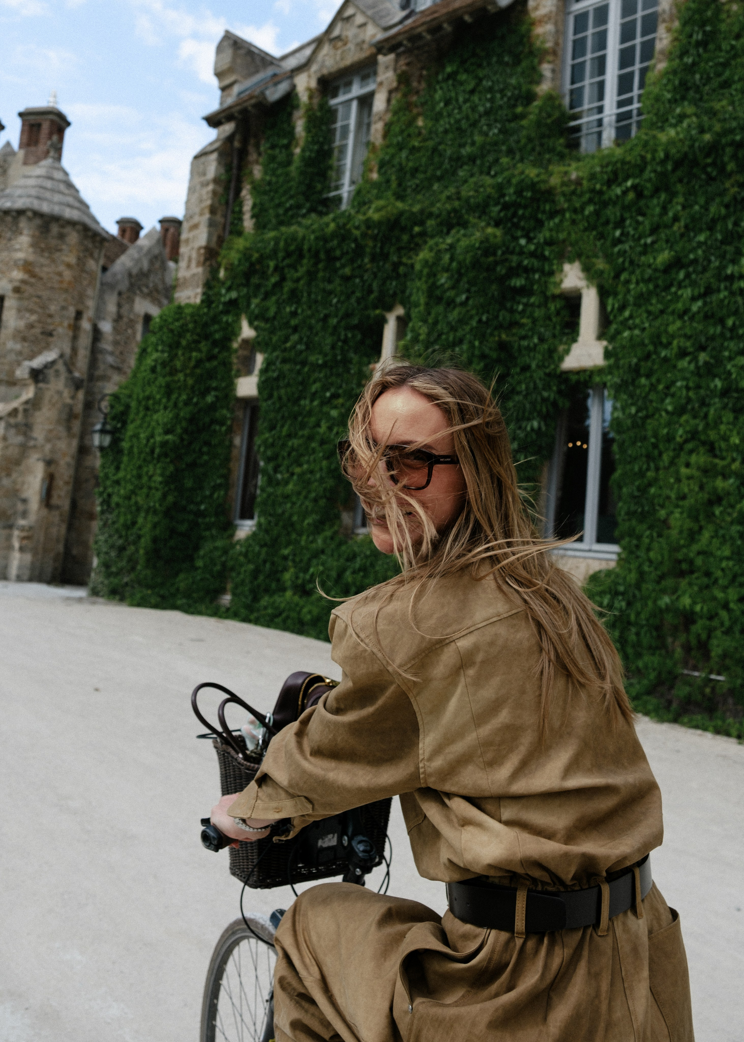 Editorial fashion portrait of a model riding a bicycle by an ivy-covered stone wall